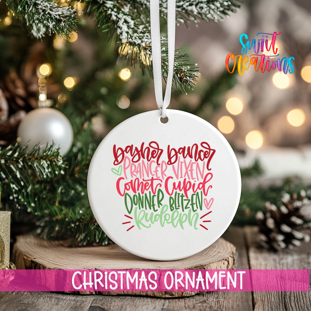 A white Christmas ornament with a colorful design hangs on a tree branch, surrounded by pine cones and other holiday decorations.
