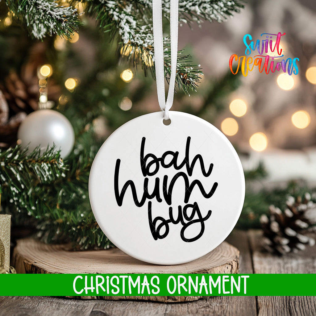 A white Christmas ornament with the text "bah hum bug" hanging from a tree branch, surrounded by pine branches and twinkling lights.