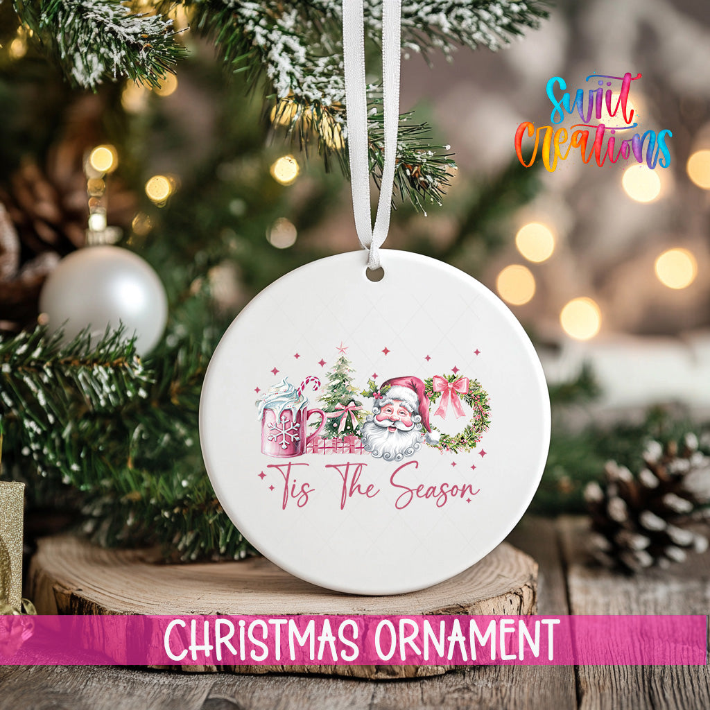A white Christmas ornament with a Santa Claus and Christmas tree design hangs on a tree branch, surrounded by other holiday decorations and lights.