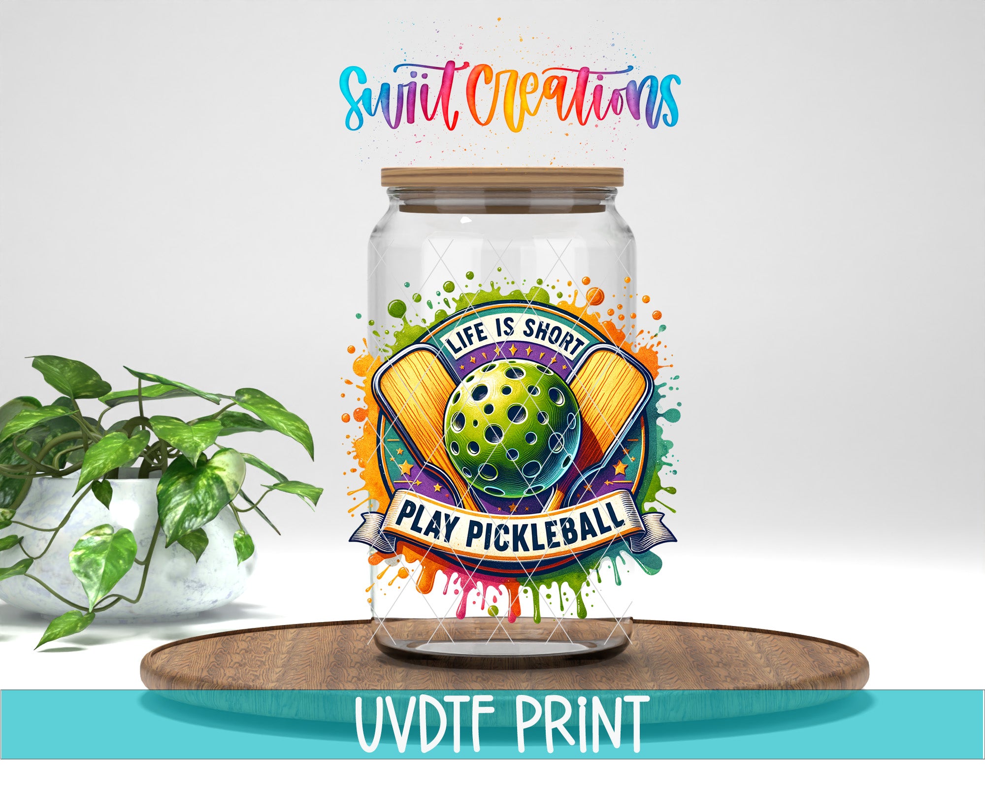 a glass jar with a colorful label that says "Life is short, play pickleball" and includes a tennis racket and a green ball. The jar is placed on a wooden surface, and there is a potted plant in the background.