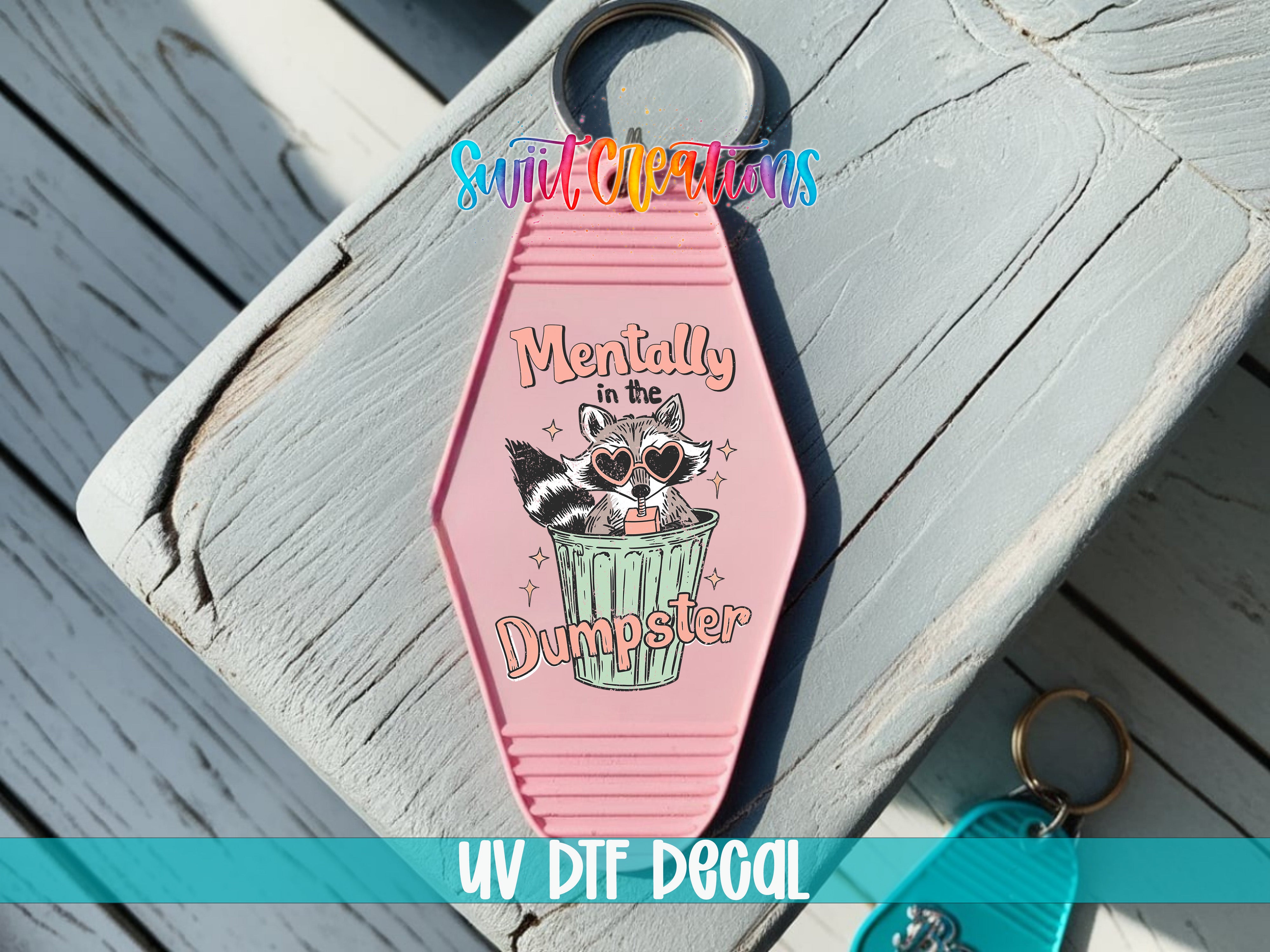 A pink keychain with a cartoon raccoon and the text "Mentally in the Dumpster" printed on it, placed on a wooden surface.