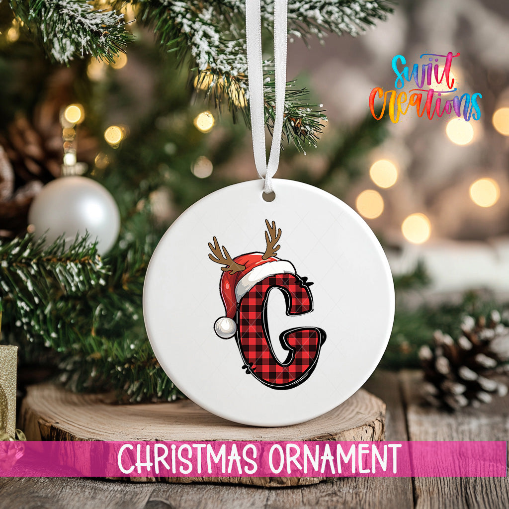 A white Christmas ornament with a red and black plaid letter "G" and a Santa hat is hanging on a tree branch.