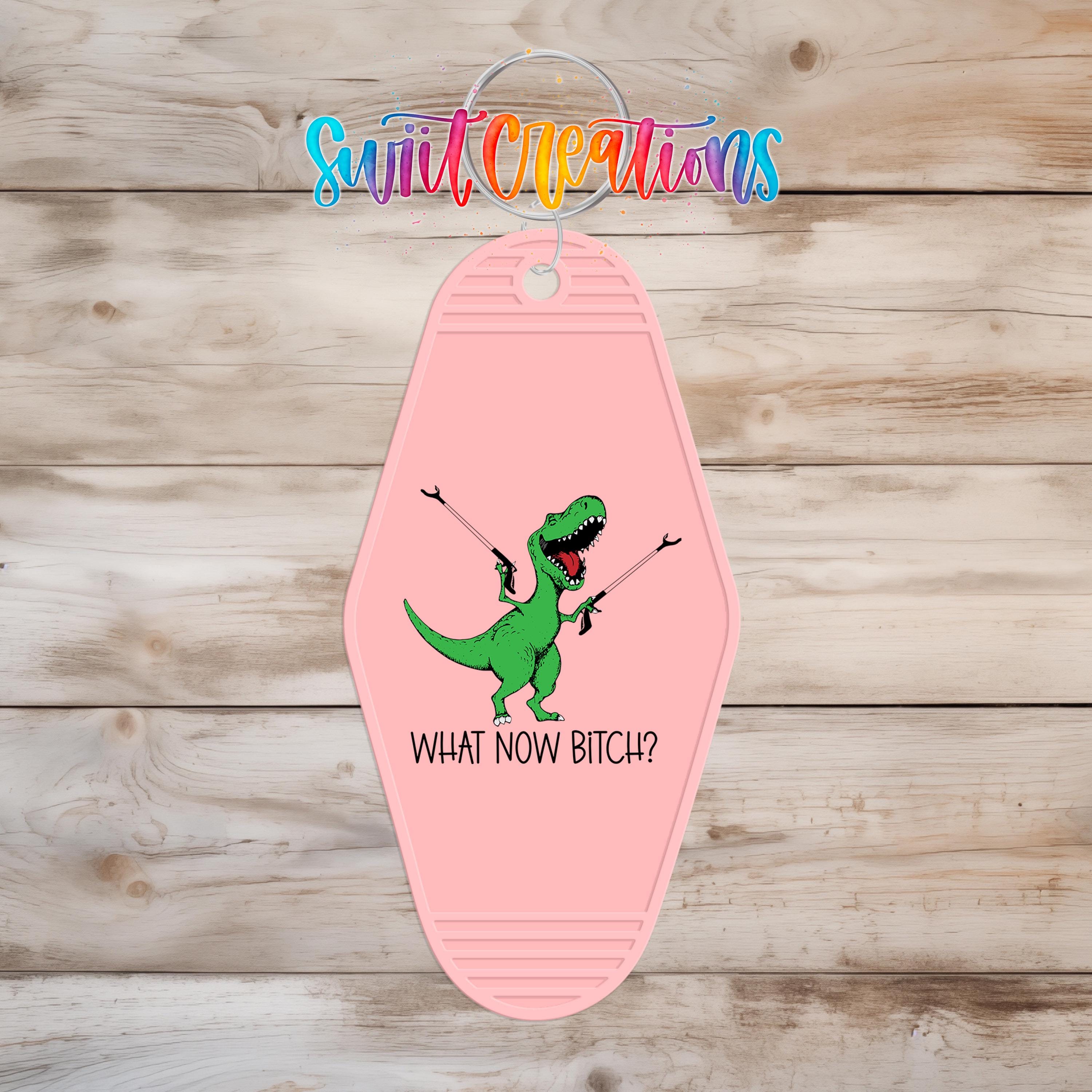 A pink keychain with a cartoon dinosaur and the text "WHAT NOW BITCH?" printed on it.