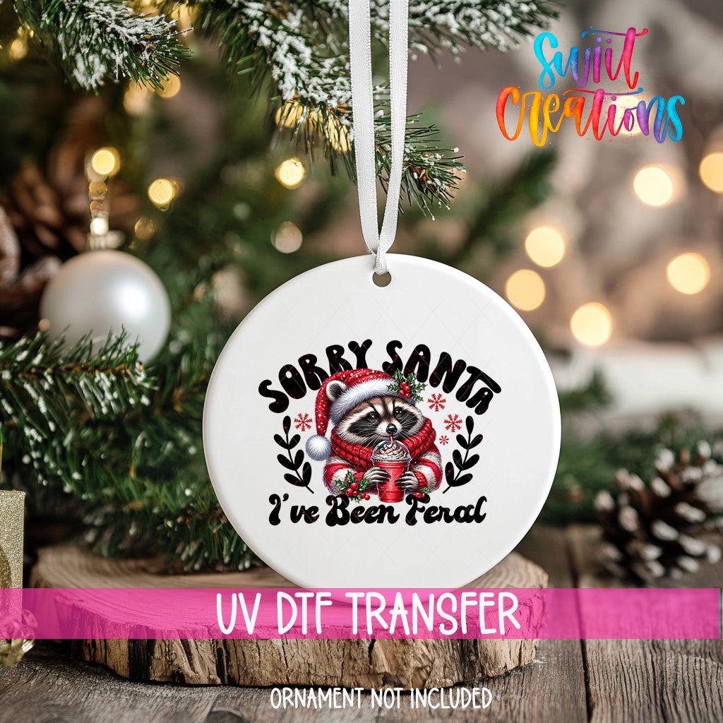 A white ornament with a raccoon wearing a Santa hat and scarf, along with the text "Sorry Santa, I've been fired" printed on it, is hanging on a Christmas tree.