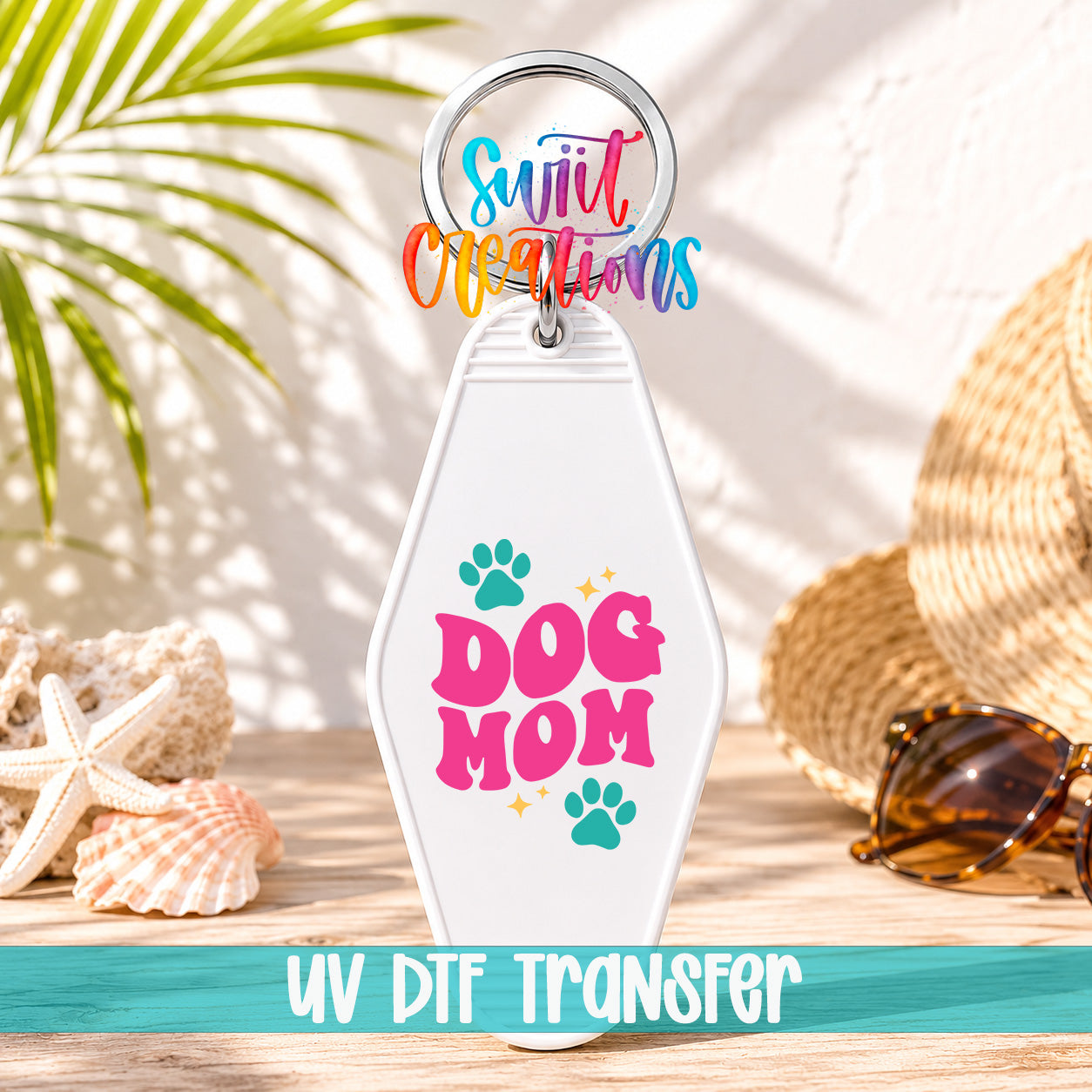 A keychain with the text "Dog Mom" printed on it, along with paw prints and a sun-like design. The keychain is placed on a wooden surface, with sunglasses and a straw hat visible in the background.
