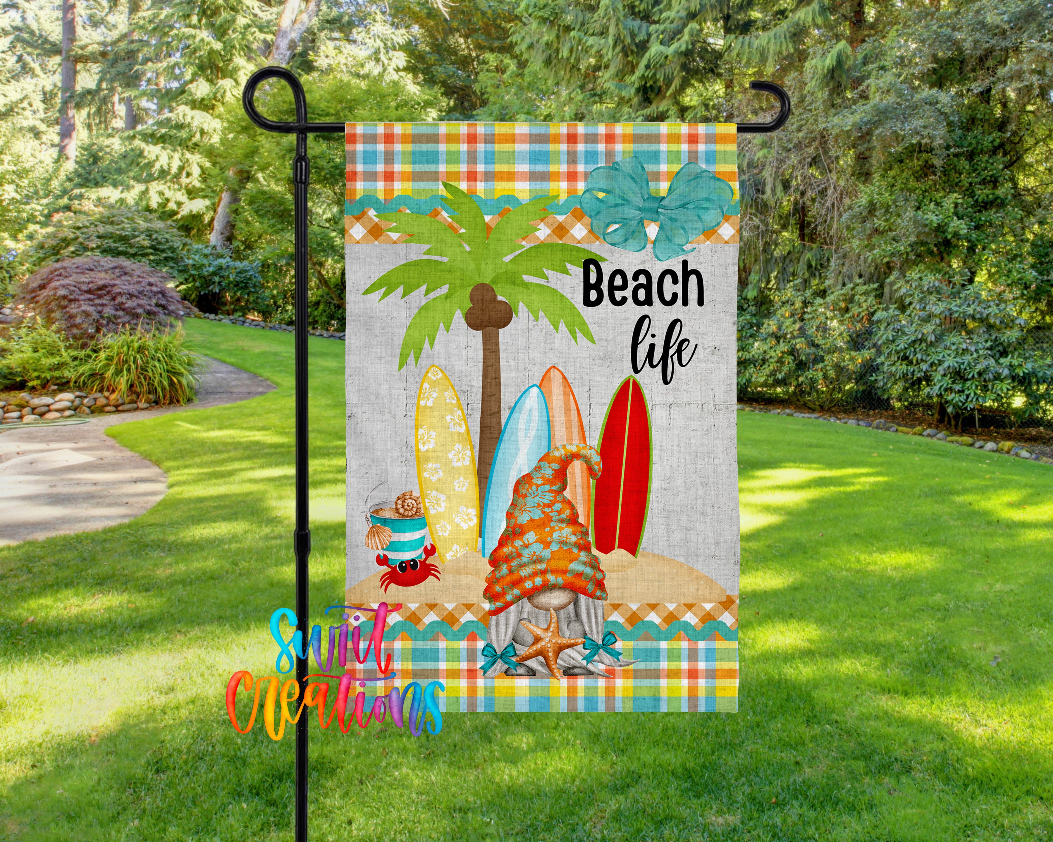 A colorful beach-themed flag with a palm tree, surfboards, and a gnome, hanging on a metal pole in a grassy area surrounded by trees.