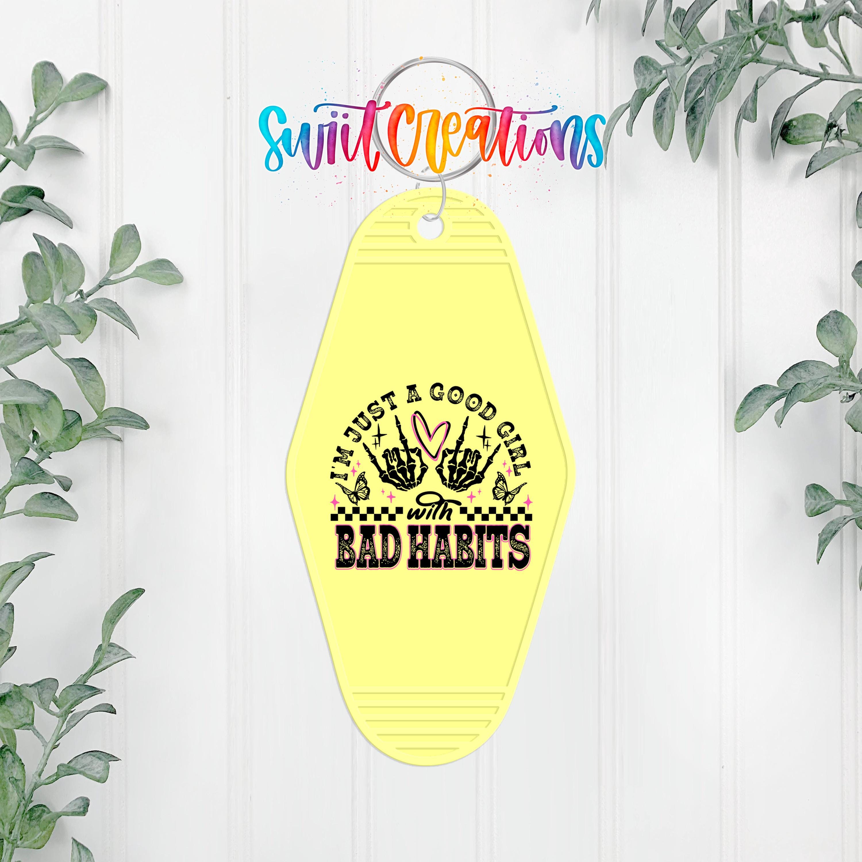 A yellow keychain with a graphic design and text, hanging on a white wall with greenery in the background.