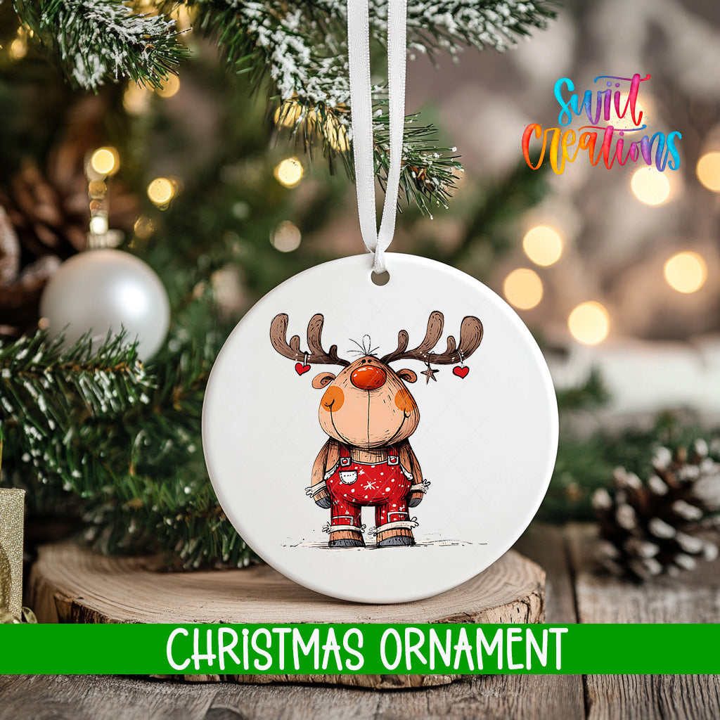 A white Christmas ornament featuring a cartoon reindeer is hanging on a tree branch, with a blurred background of lights and greenery.
