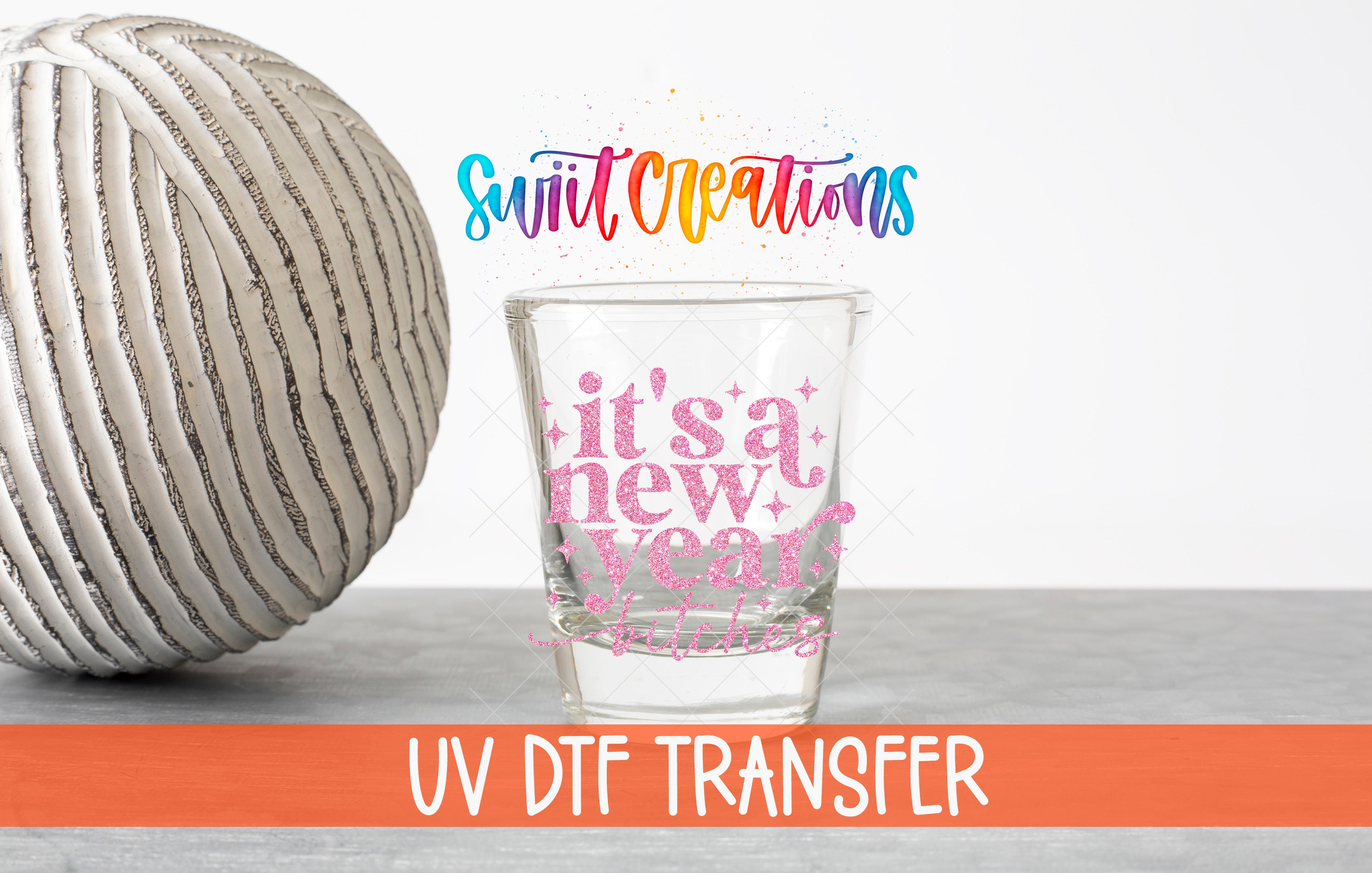 a glass with a colorful design and text, placed next to a white ball with a striped pattern. The text on the glass reads "it's a new year" and "UV DFT Transfer".