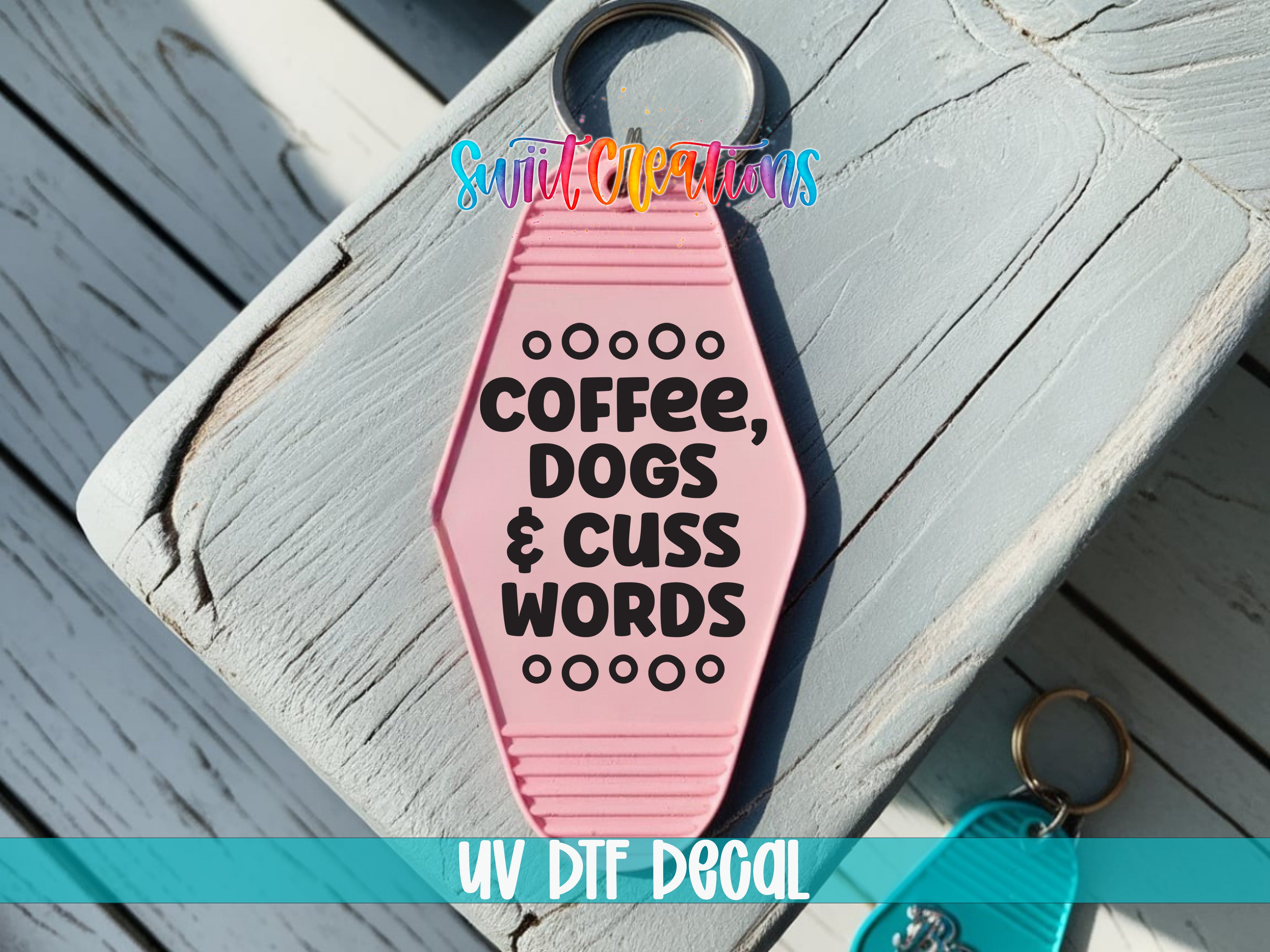 A pink keychain with the text "COFFEE, DOGS & CUSS WORDS" printed on it, placed on a wooden surface.