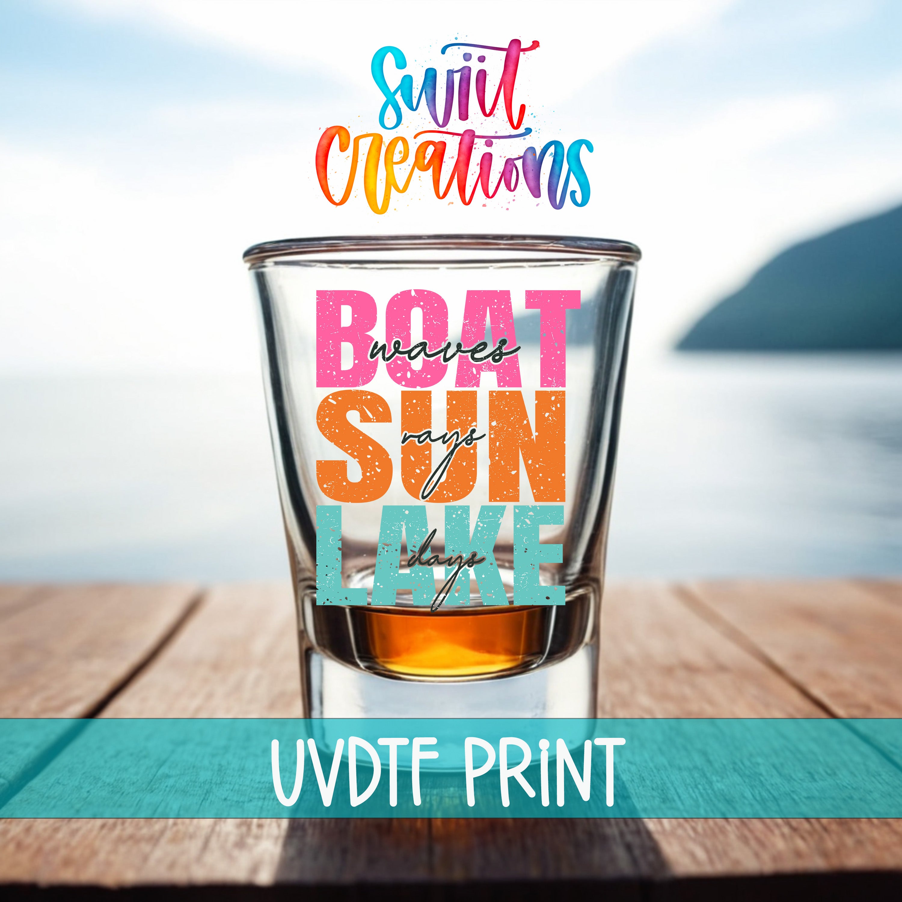 a colorful glass with text on it, placed on a wooden surface with a blurred background that includes a body of water and a mountain.