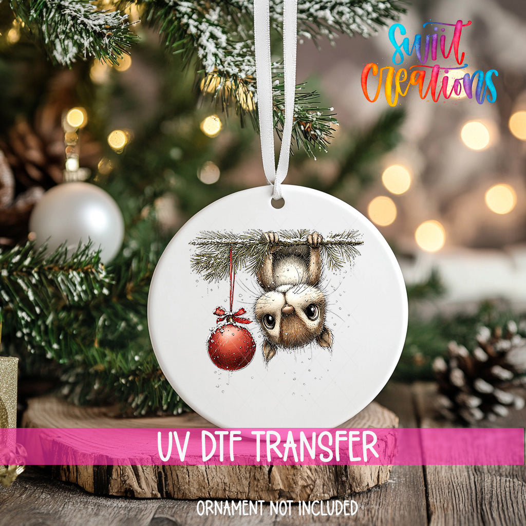 A white ceramic ornament with a cute illustration of a cat hanging from a branch, holding a red ornament, is displayed on a Christmas tree.