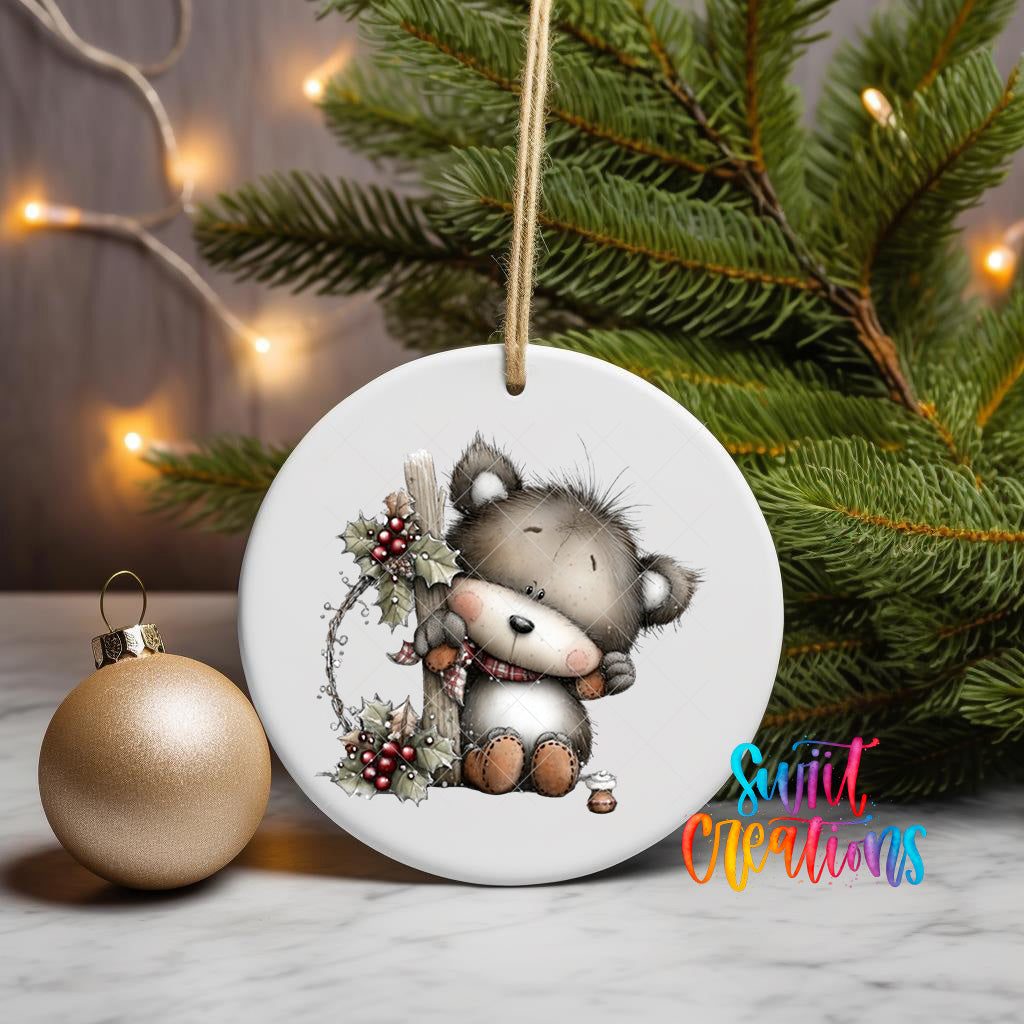 A white ceramic ornament featuring a cute teddy bear is displayed on a table, with a gold ornament and a Christmas tree in the background.