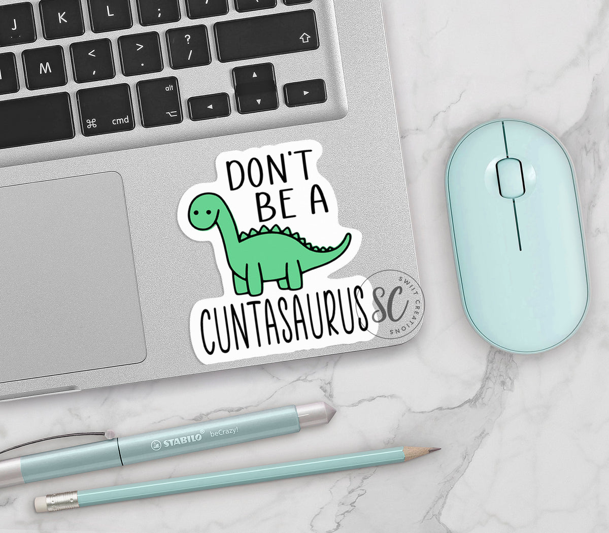 Don't Be a C*ntasaurus vinyl sticker on a silver laptop next to a mint mouse and pens