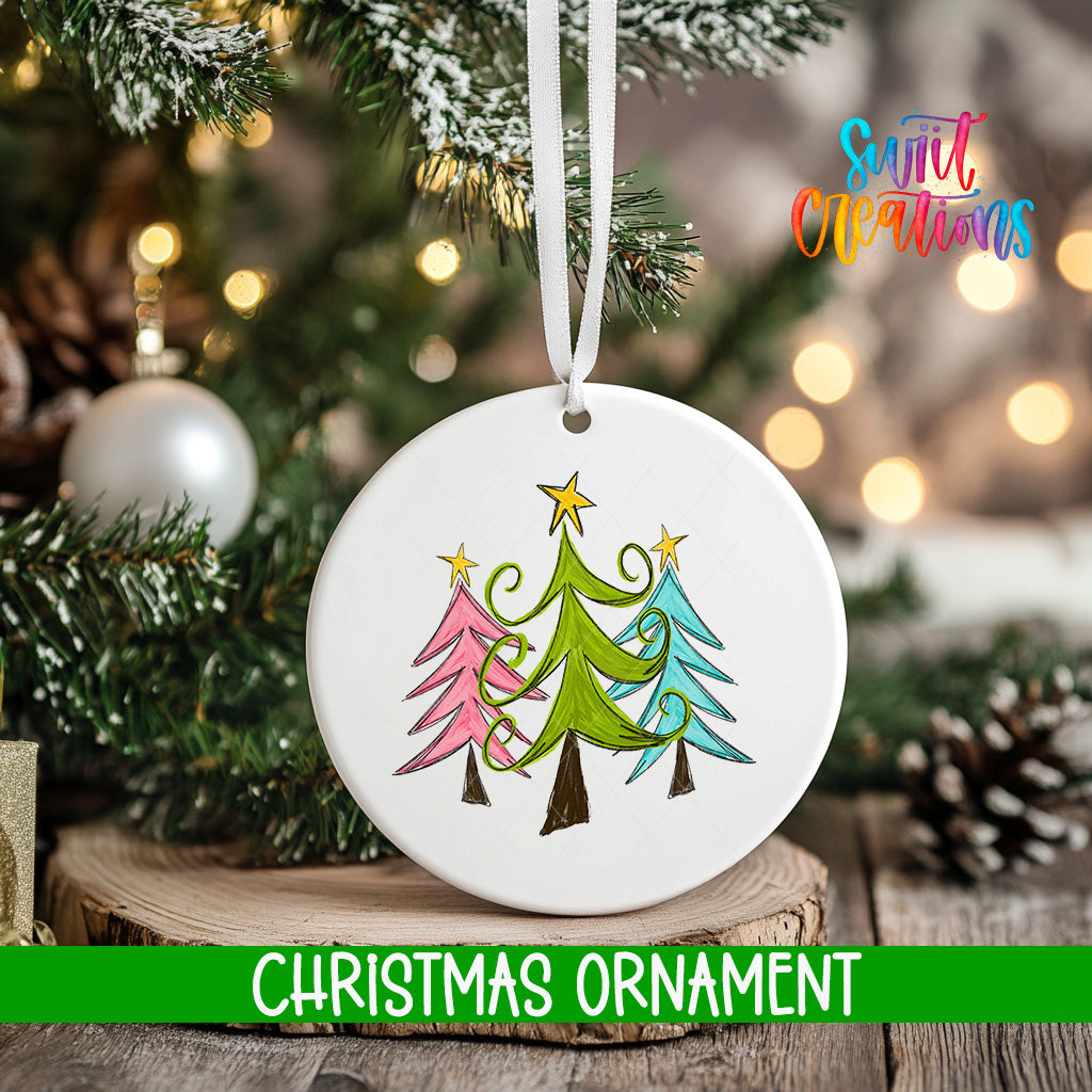 A white Christmas ornament with a colorful illustration of three trees hangs on a tree branch, surrounded by twinkling lights and greenery.