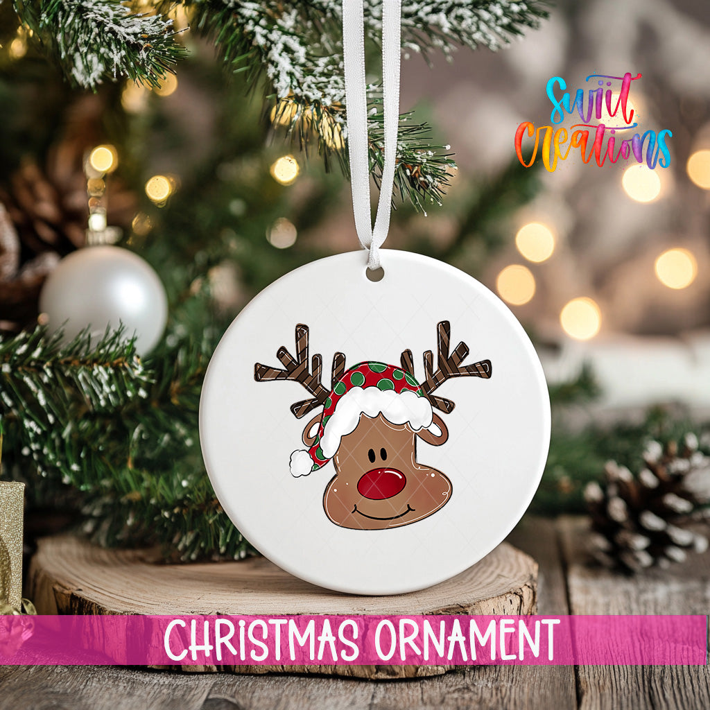 A white Christmas ornament with a reindeer face and a Santa hat is hanging on a tree branch, with a blurred background of lights and pine cones.