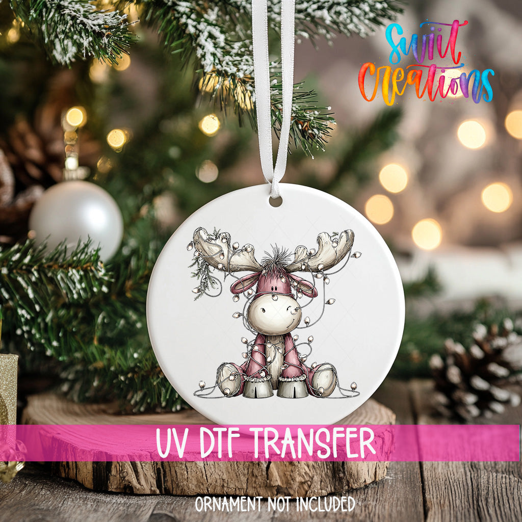 A white ornament with a deer-like figure is hanging on a Christmas tree, with a blurred background of pine branches and twinkling lights.