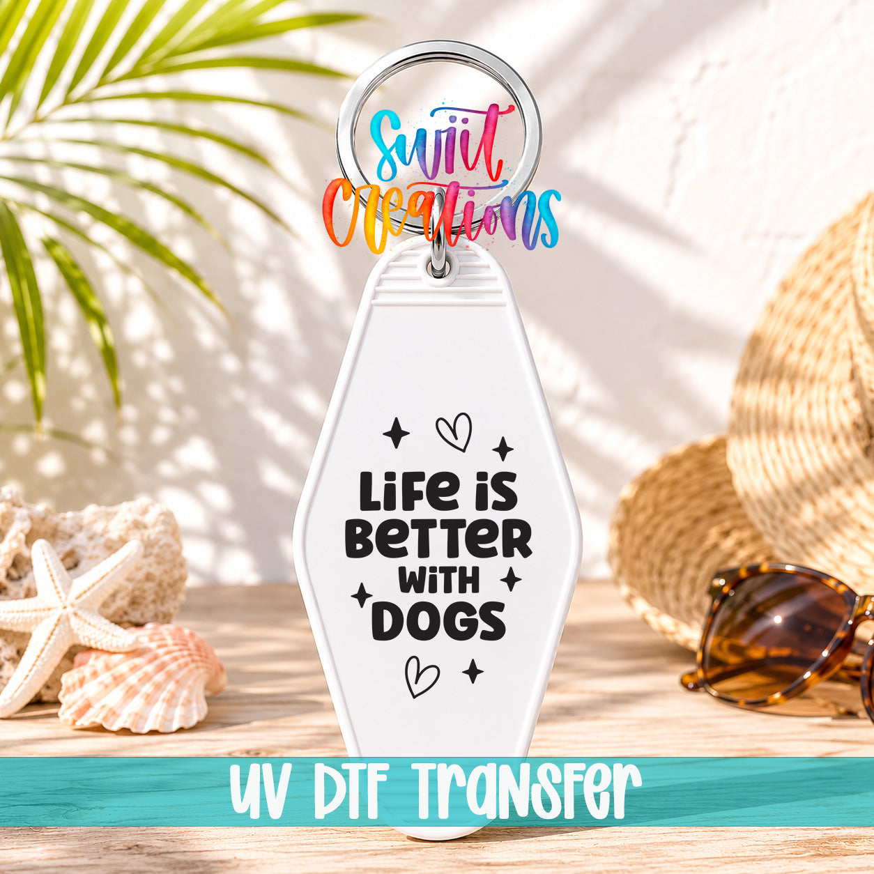 A keychain with the text "Life is better with dogs" printed on it, along with a colorful ring and a straw hat. The keychain is placed on a wooden surface, with sunglasses and a palm frond visible in the background.