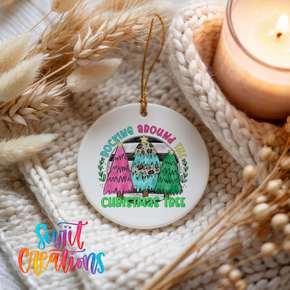 A white ceramic ornament with a colorful design hangs on a white knitted blanket, surrounded by dried wheat and a lit candle.