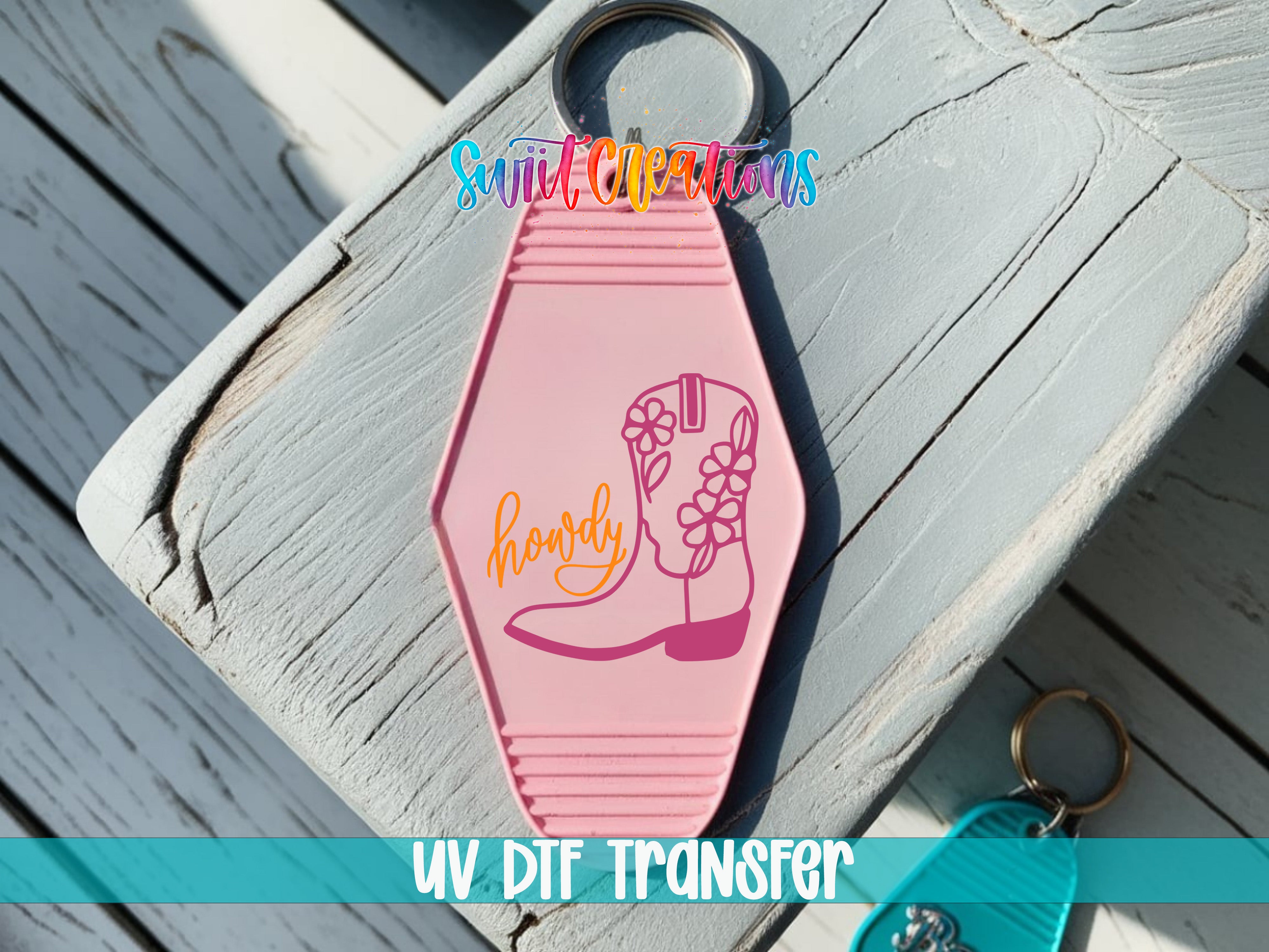A pink keychain with a cowboy boot design and the word "howdy" printed on it, placed on a wooden surface.