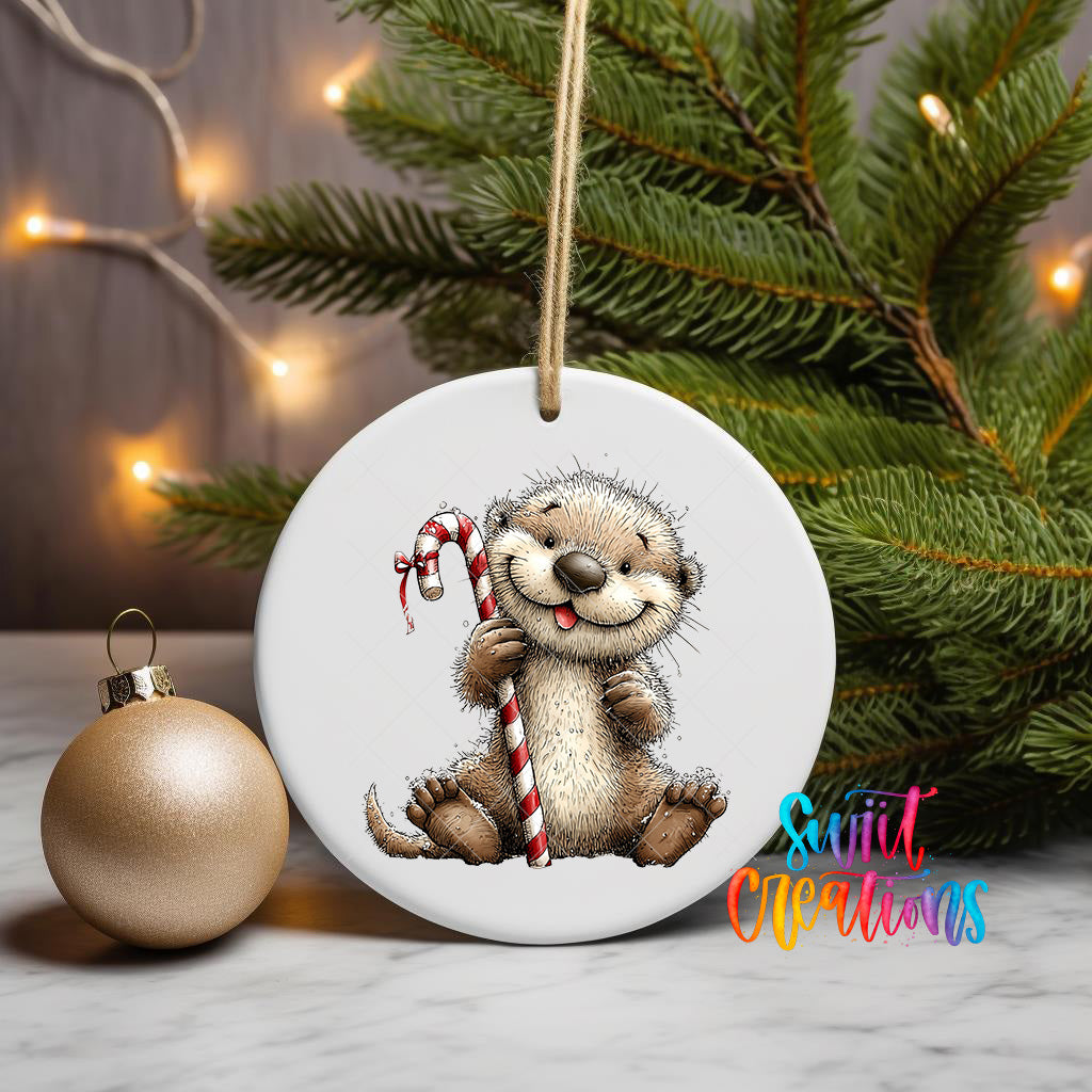 A white ceramic ornament featuring a smiling otter holding a candy cane, placed on a marble surface next to a gold ornament and a Christmas tree with twinkling lights.