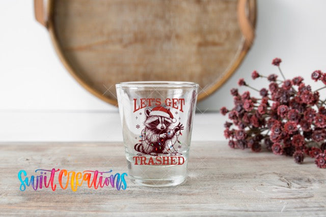 A shot glass with a raccoon wearing a Santa hat and the text "Let's get trashhed" printed on it, placed on a wooden surface next to a bunch of red flowers.