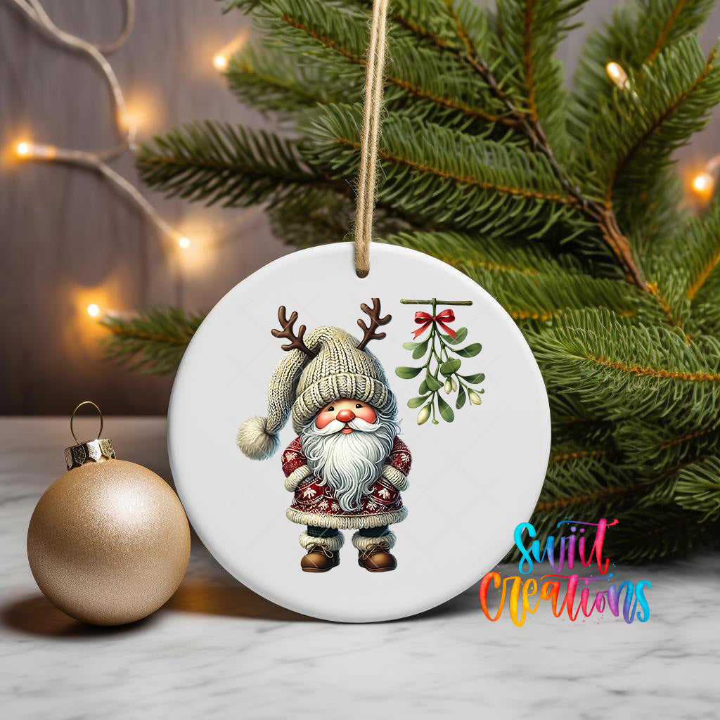 A white ornament featuring a gnome with antlers and a mistletoe is displayed on a white surface, with a Christmas tree and twinkling lights in the background.