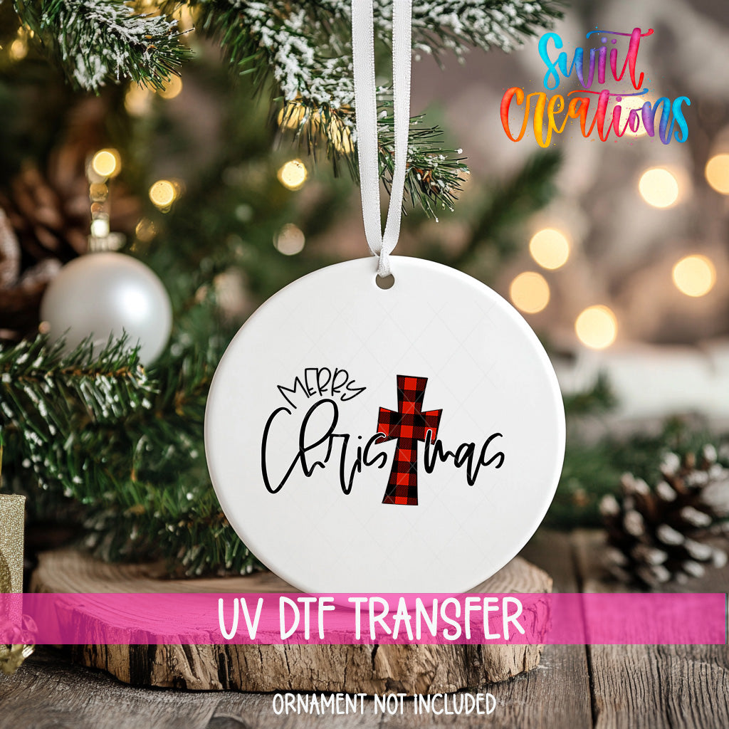 A white Christmas ornament with a red and black plaid cross design hangs on a tree branch, surrounded by pine branches and twinkling lights.