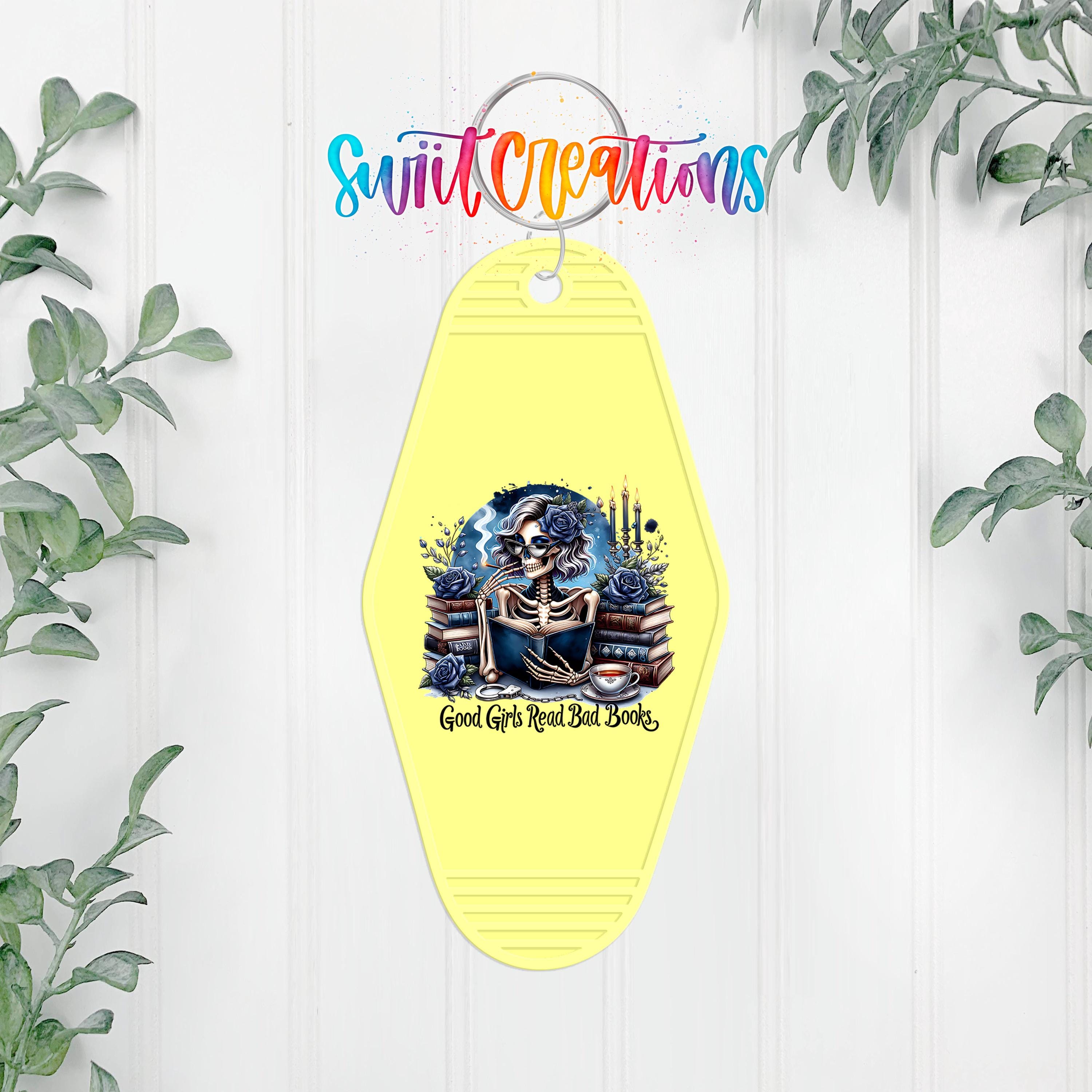 A yellow keychain with a skull and crossbones design, surrounded by greenery, and the text "Good Girls Read Bad Books" printed on it.