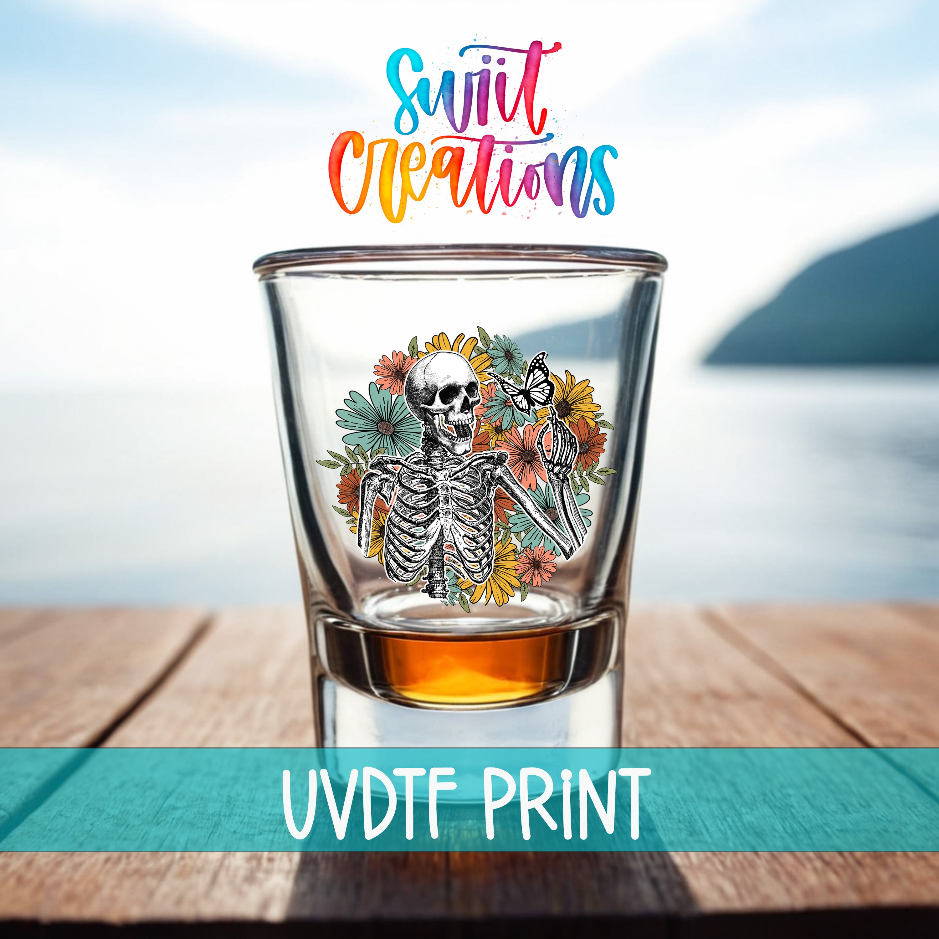 A shot glass with a skeleton design and colorful flowers, placed on a wooden surface with a body of water in the background.