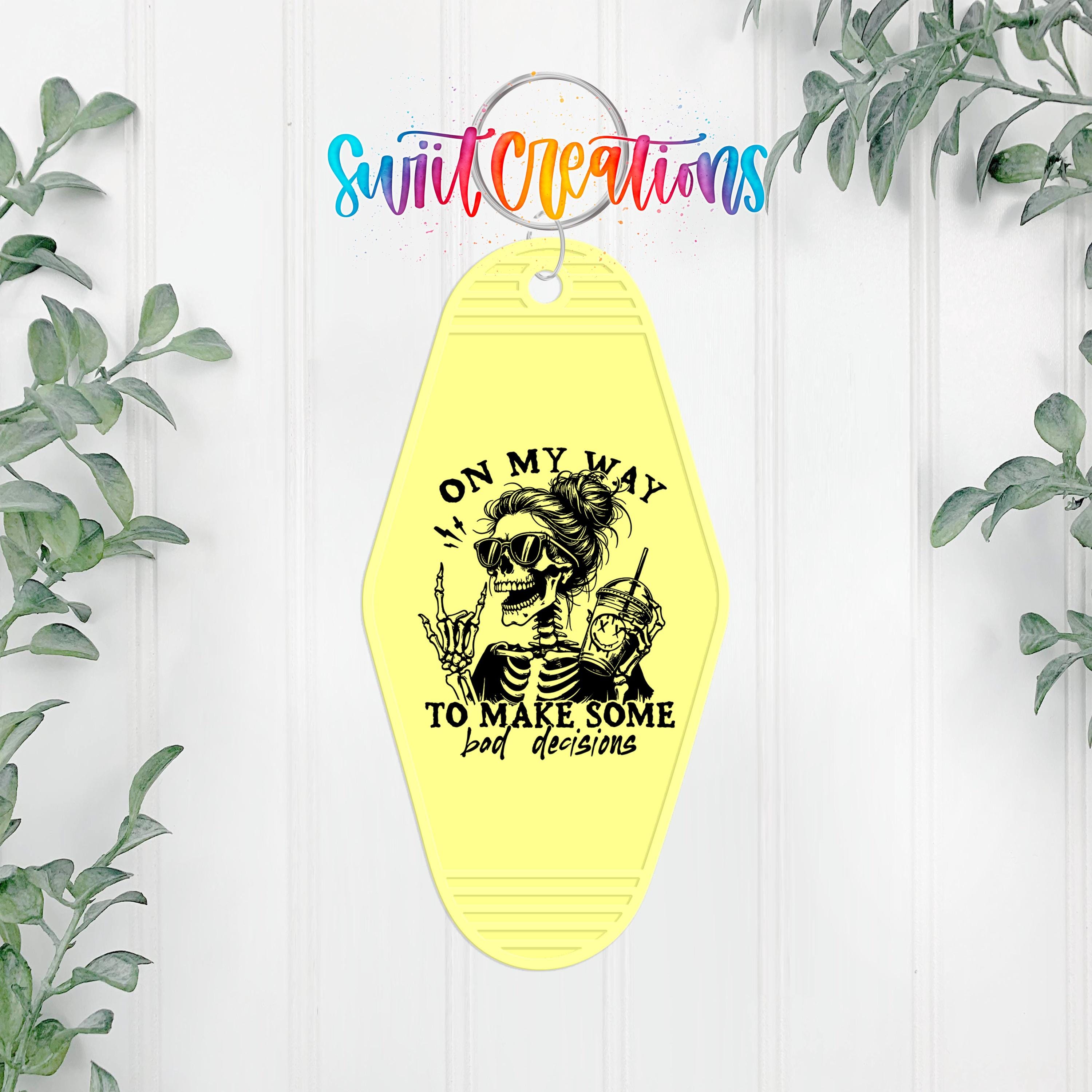 A yellow keychain with a skeleton graphic and the text "I ON MY WAY TO MAKE SOME BAD DECISIONS" printed on it, hanging on a white wall with greenery in the background.