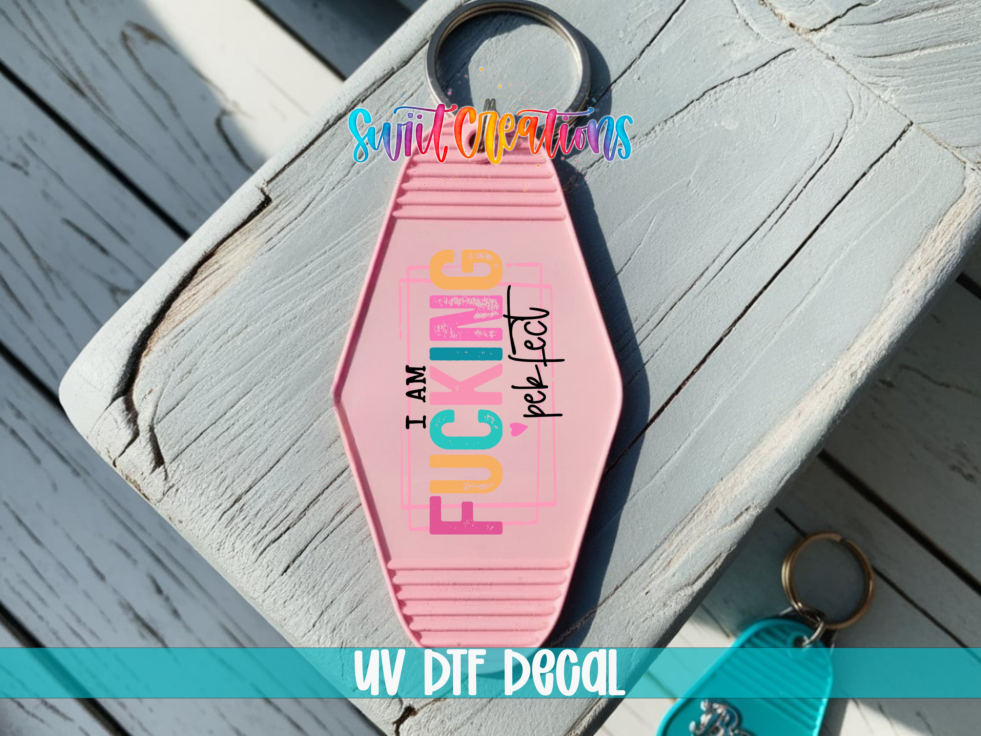 A pink keychain with the text "I AM FUCKING PERFECT" printed on it, placed on a wooden surface.
