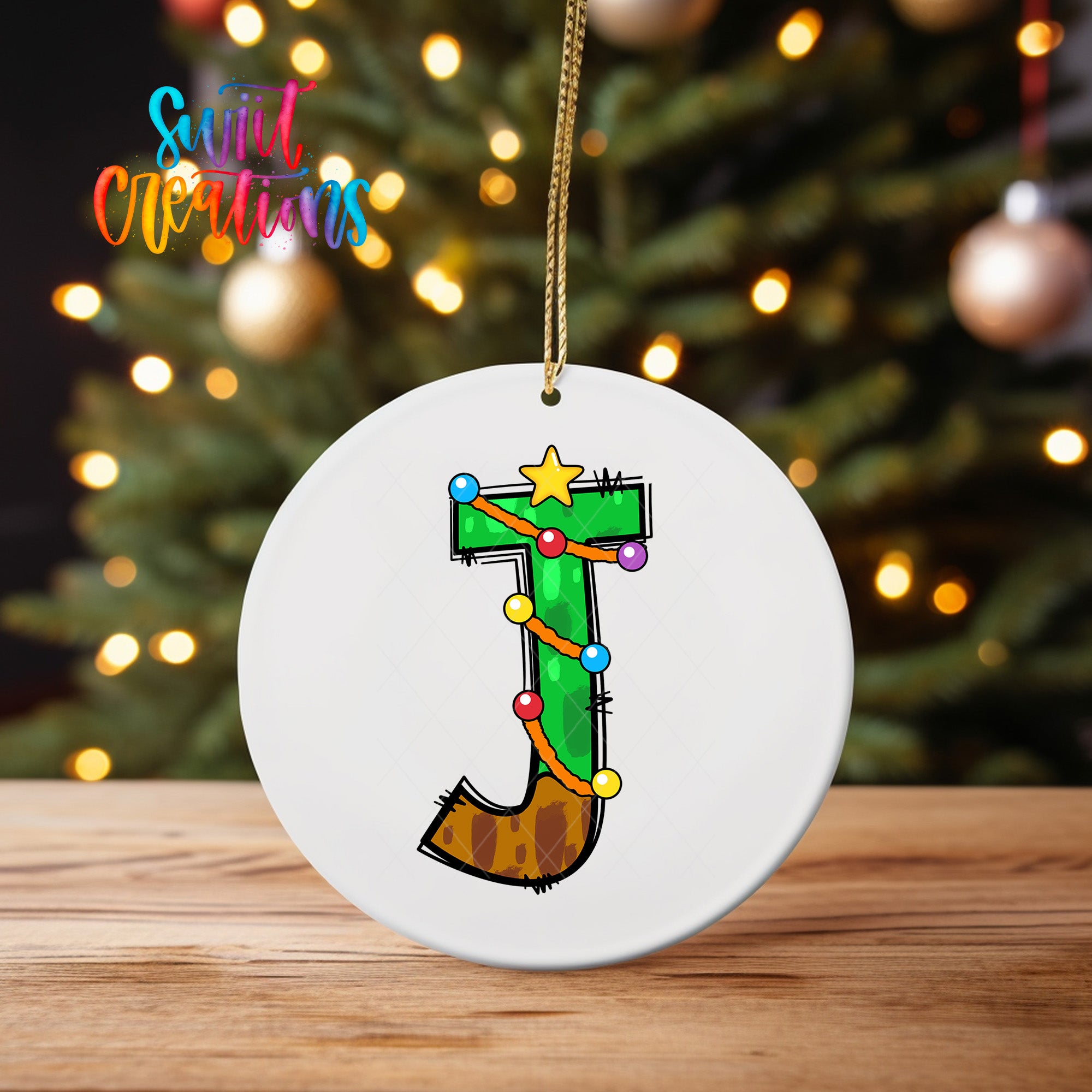 A white ornament with a colorful design featuring the letter "J" and Christmas lights is displayed on a wooden surface.