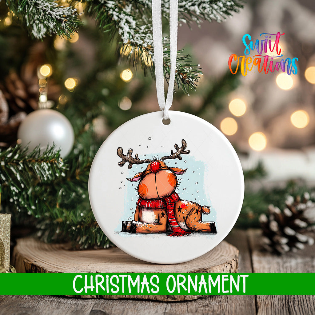 A white Christmas ornament featuring a cartoon reindeer with antlers and a red scarf, hanging on a tree branch with a blurred background of lights.