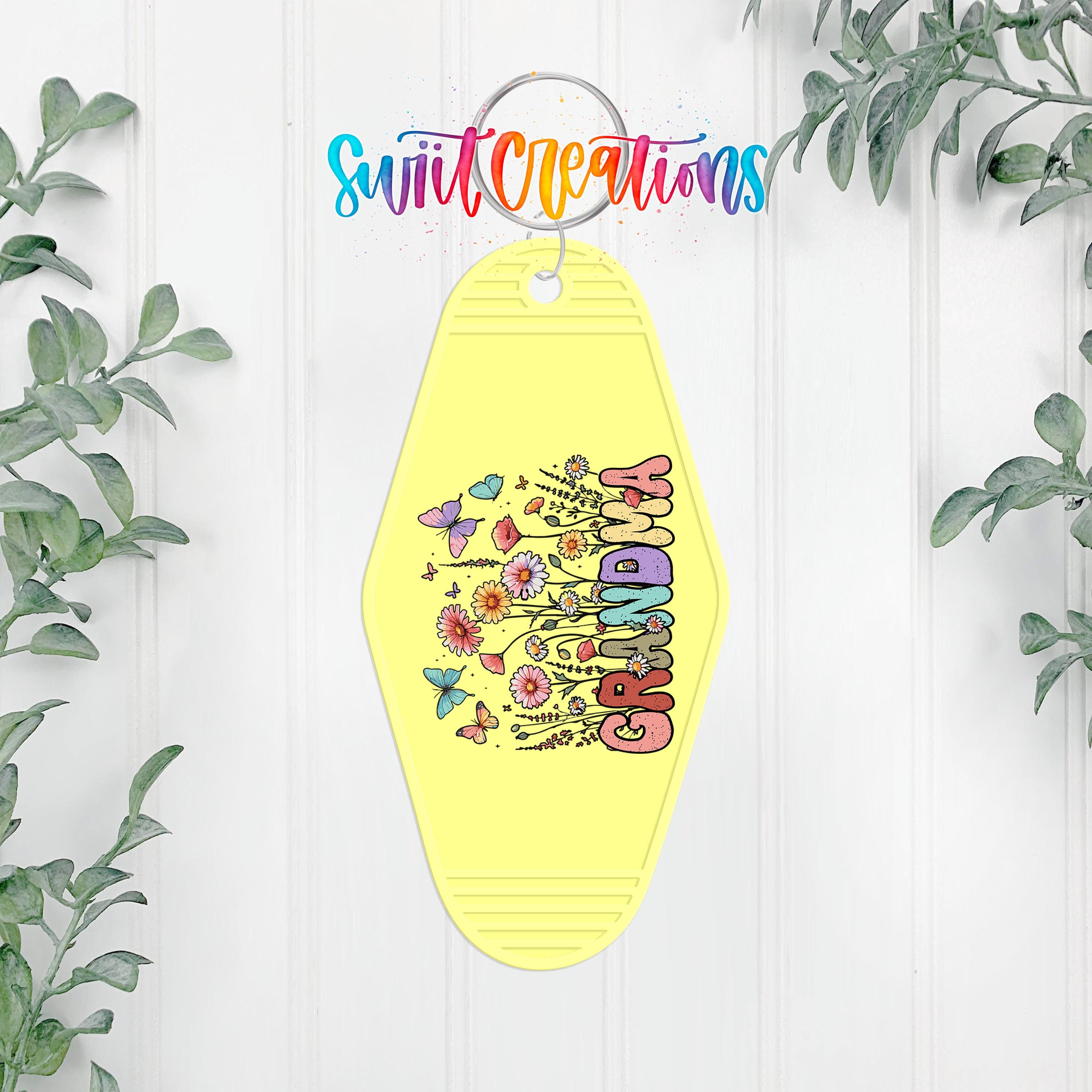 A yellow keychain with a colorful design and the word "GRANDMA" printed on it, hanging on a white wall with greenery in the background.