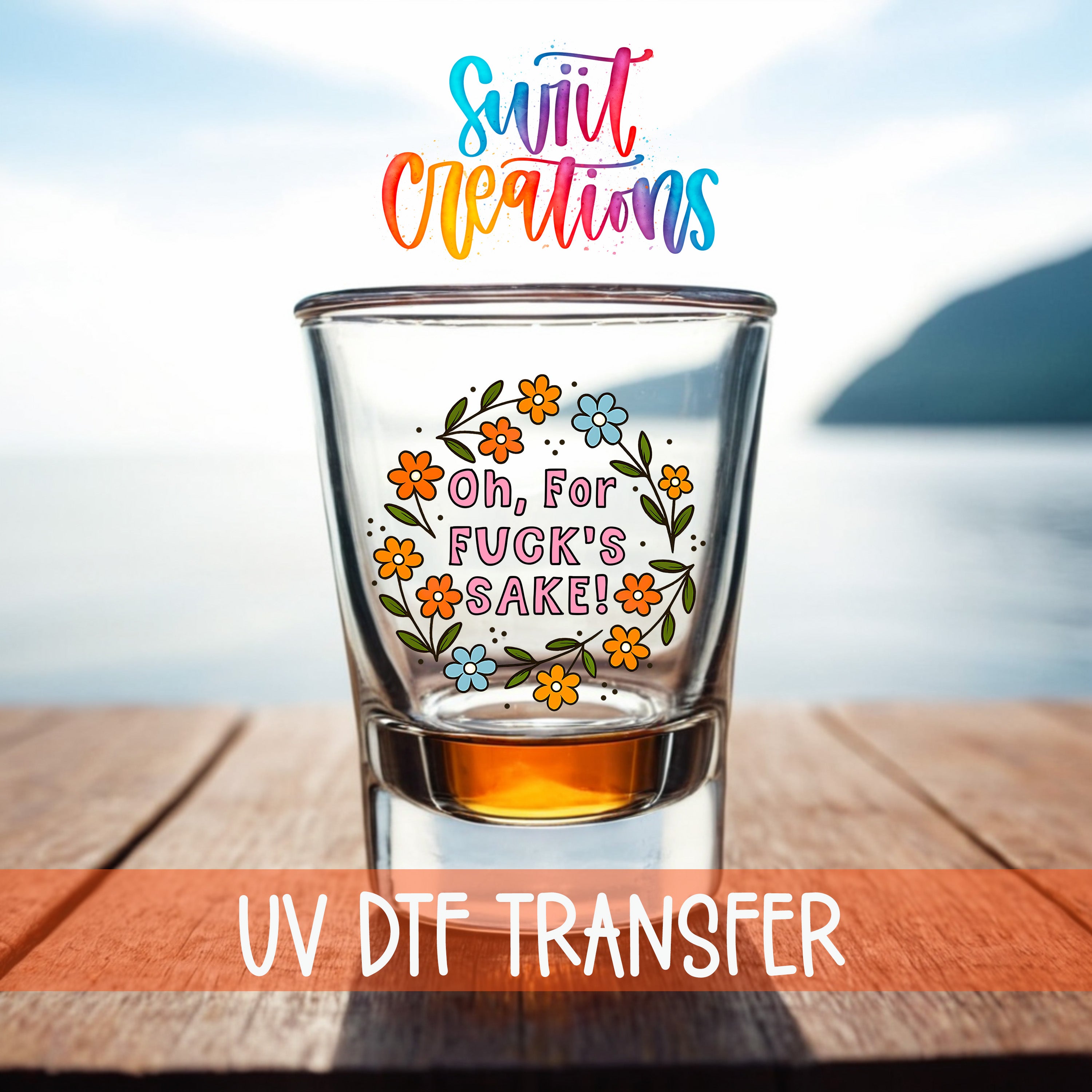 A shot glass with a colorful floral design and text on it, placed on a wooden surface with a body of water in the background.