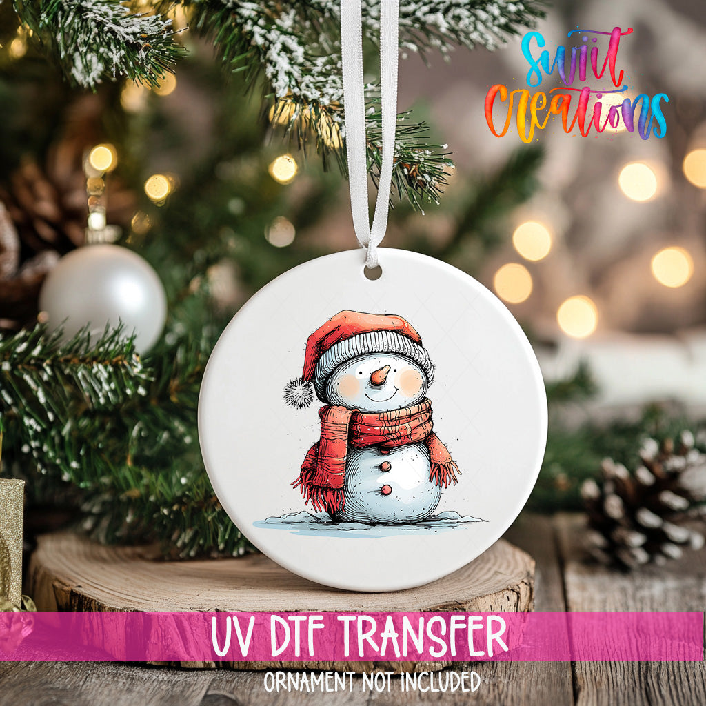 A white ornament with a snowman design hangs on a Christmas tree, surrounded by pine branches and twinkling lights.