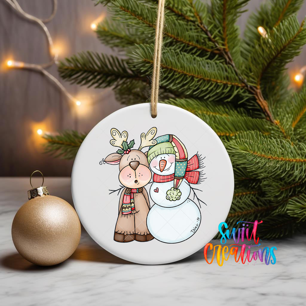 A white ceramic ornament featuring a cartoon illustration of a reindeer and a snowman, with a Christmas tree in the background and a gold ornament to the left.
