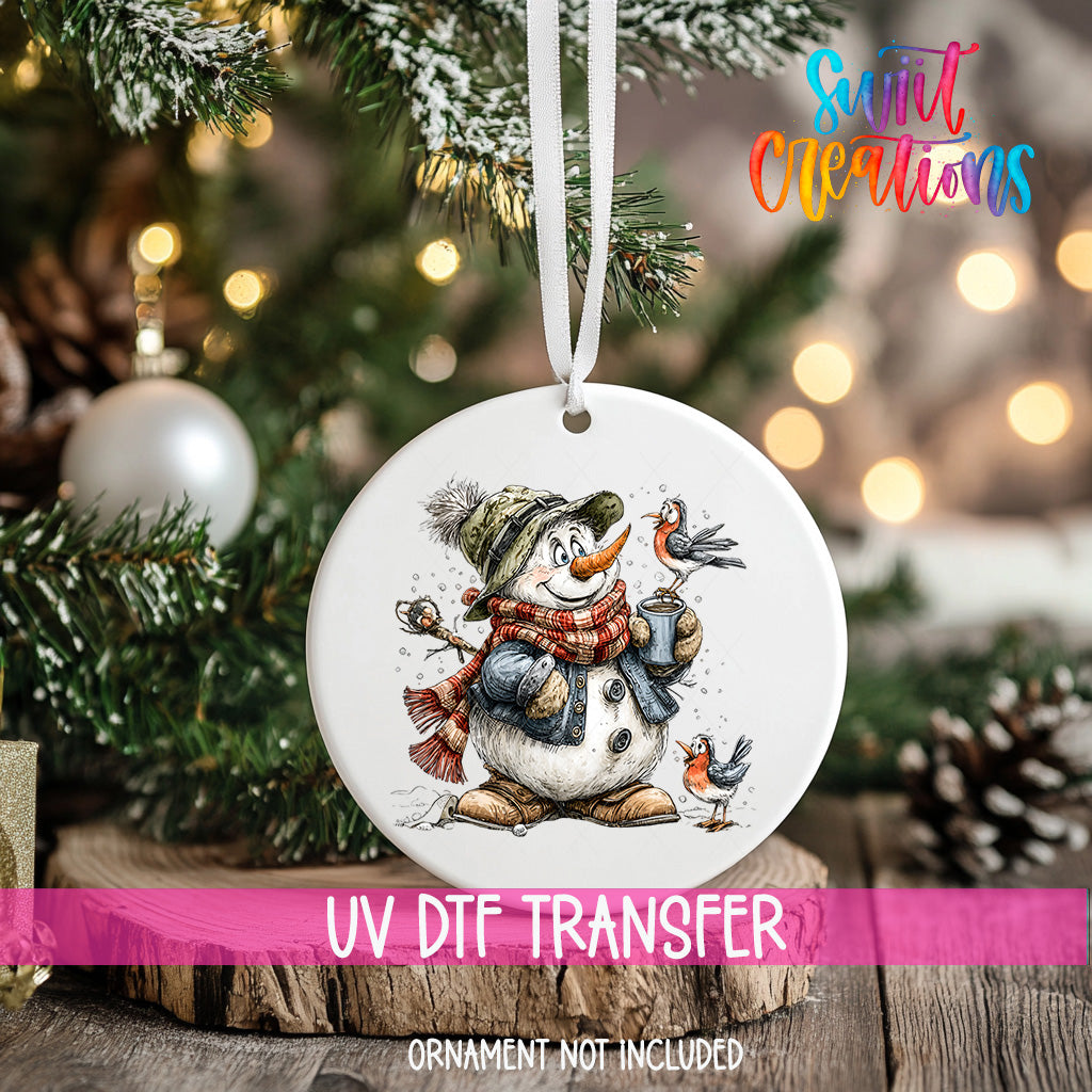 A white ornament with a snowman and birds is hanging on a Christmas tree, with a pink ribbon around it.