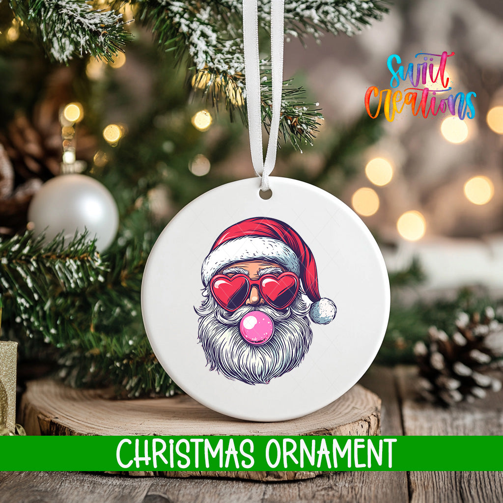 A white Christmas ornament with a Santa Claus face wearing sunglasses and a red hat, with a pink bubble gum bubble in his mouth, is hanging from a tree branch.