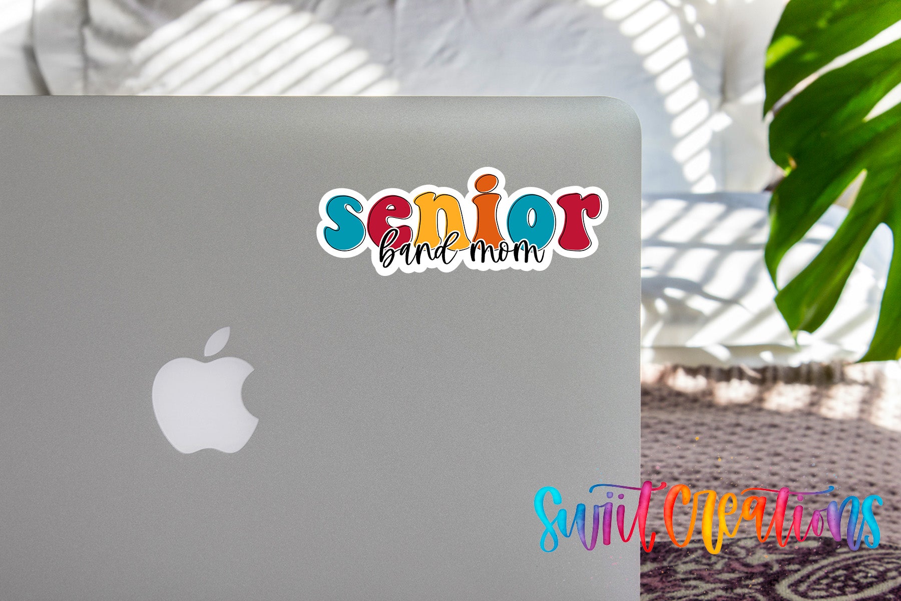 a sticker with the text "senior band mom" on the back of a silver laptop, with a plant and a patterned rug visible in the background.