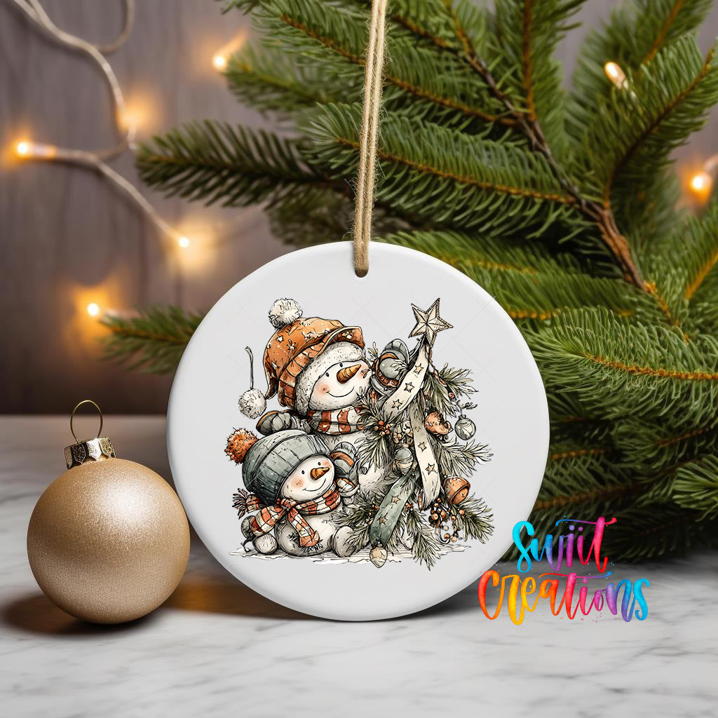 A white ceramic ornament featuring a snowman and a star, hanging on a string, is displayed next to a gold ornament and a Christmas tree.