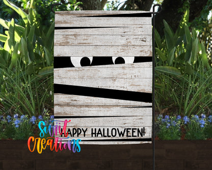 A Halloween-themed garden sign with a wooden board and a pair of white eyes peeking out from behind a black line, surrounded by lush greenery and colorful flowers.