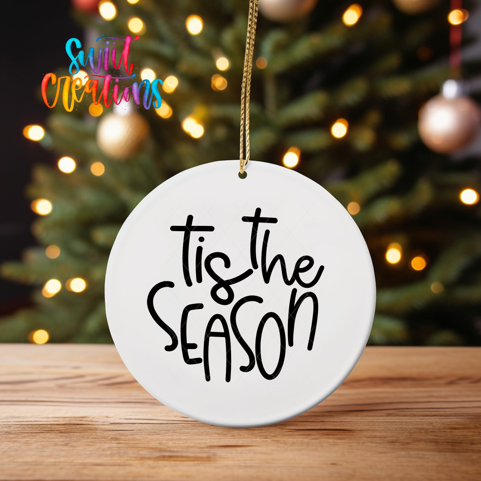 A white ceramic ornament with the text "Tis the SEASON" written on it, hanging from a string. The ornament is placed on a wooden surface, with a blurred background of a Christmas tree and colorful lights.