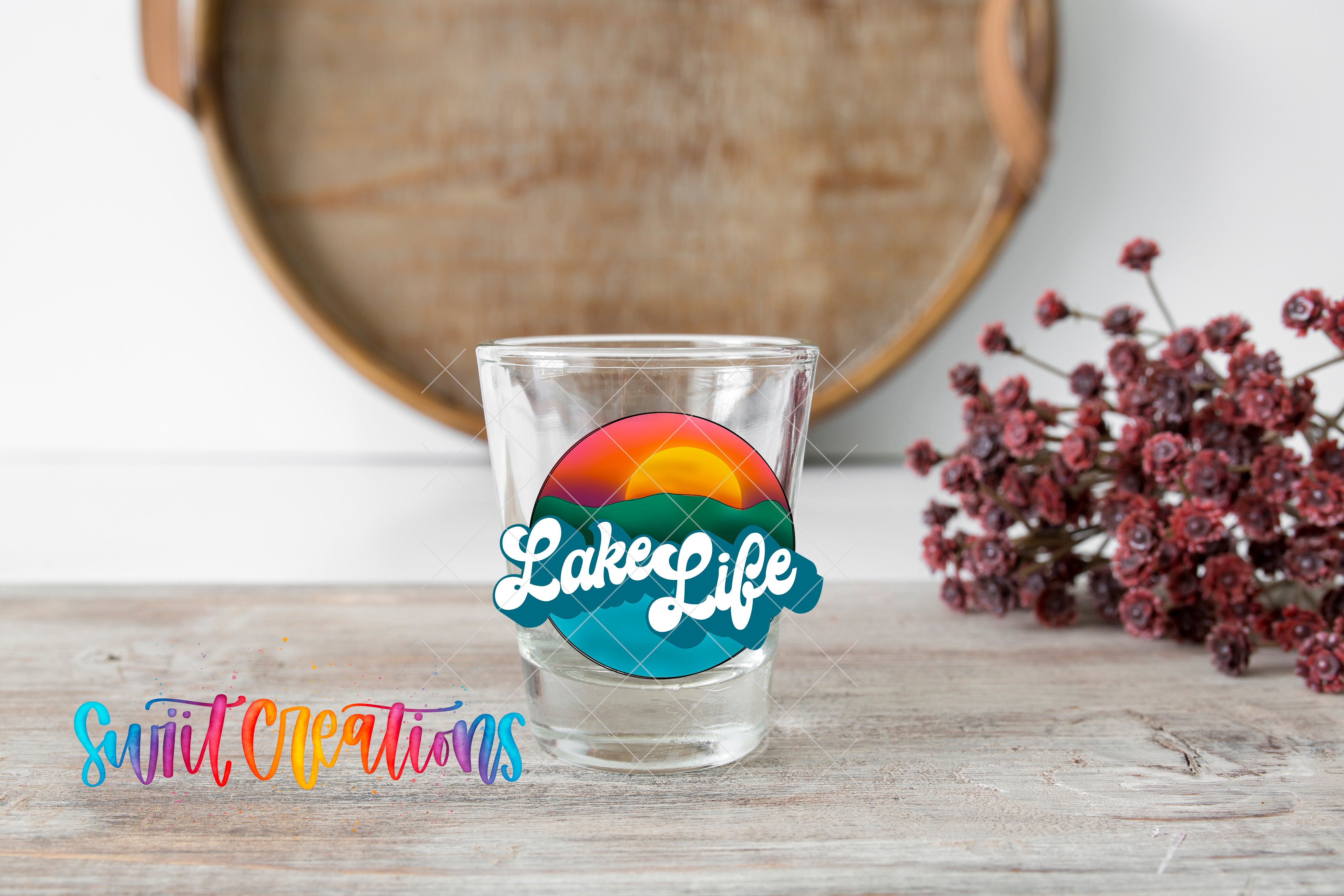 A shot glass with a colorful graphic and the text "LakeLife" on it, placed on a wooden surface next to a bunch of red flowers.