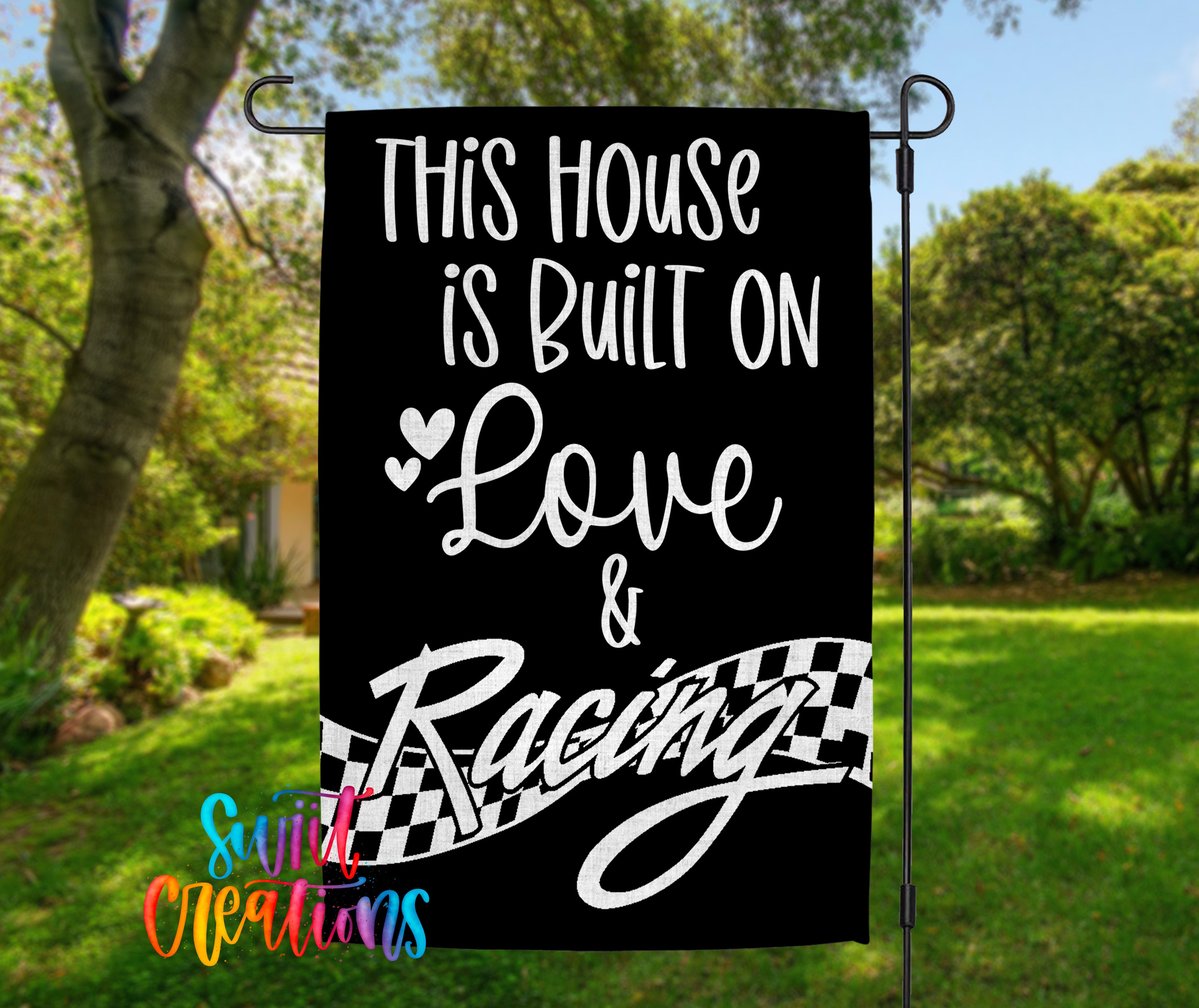 a black and white sign with white text that reads "This house is built on love & racing", placed in a grassy area with trees in the background.
