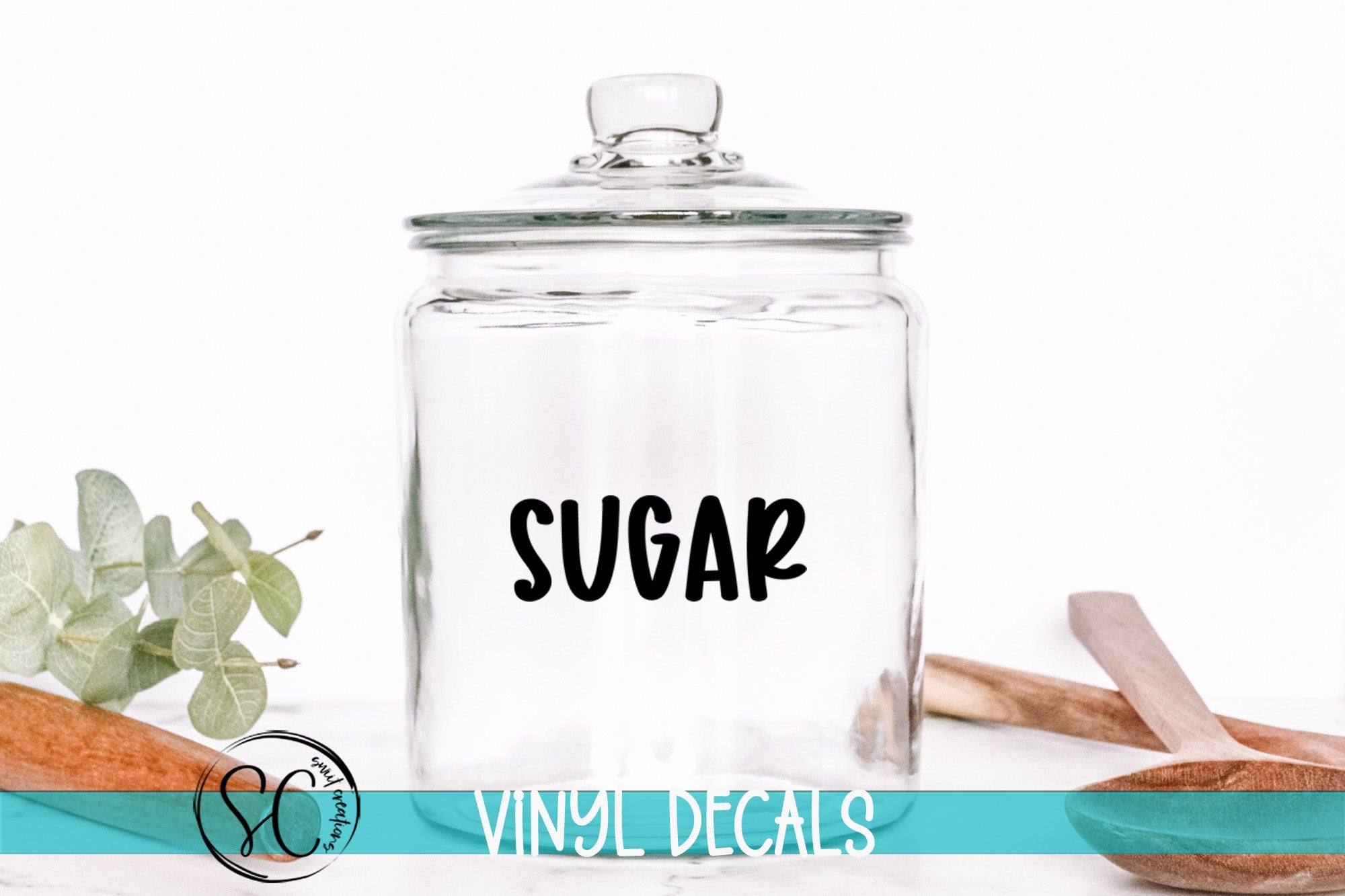 A clear glass jar with the word "SUGAR" printed on it, placed on a white surface next to some wooden utensils.