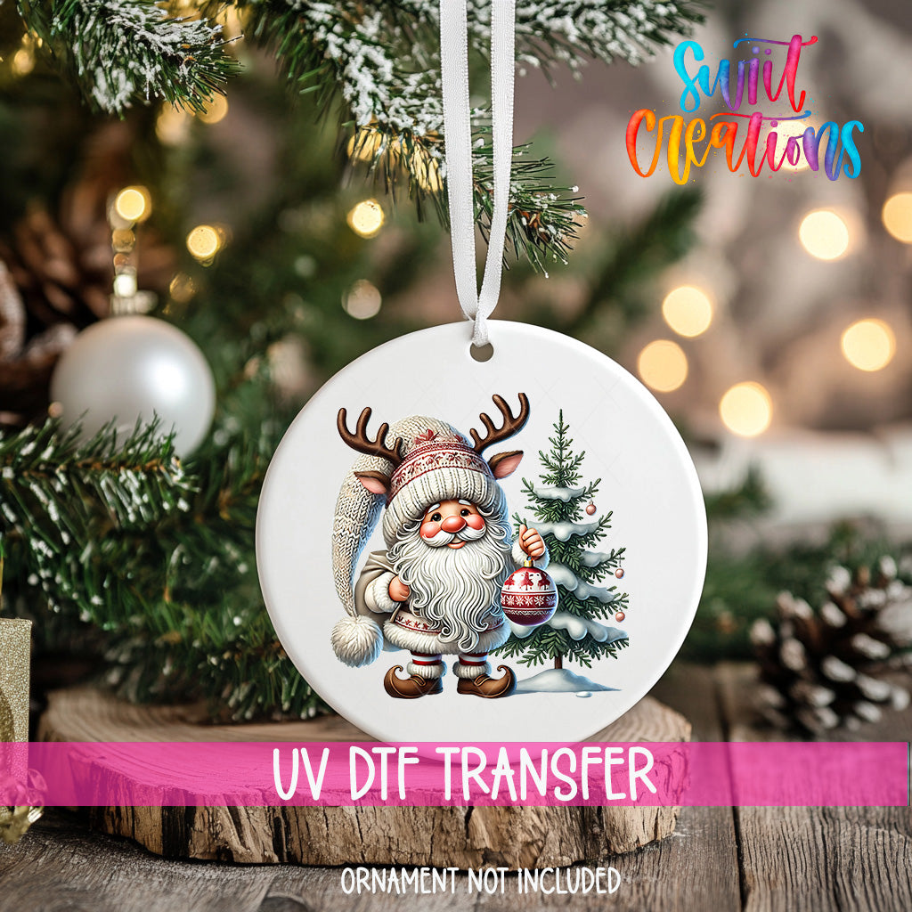A white ornament with a Santa Claus figure and a Christmas tree is hanging on a tree branch, with a pink ribbon around it.