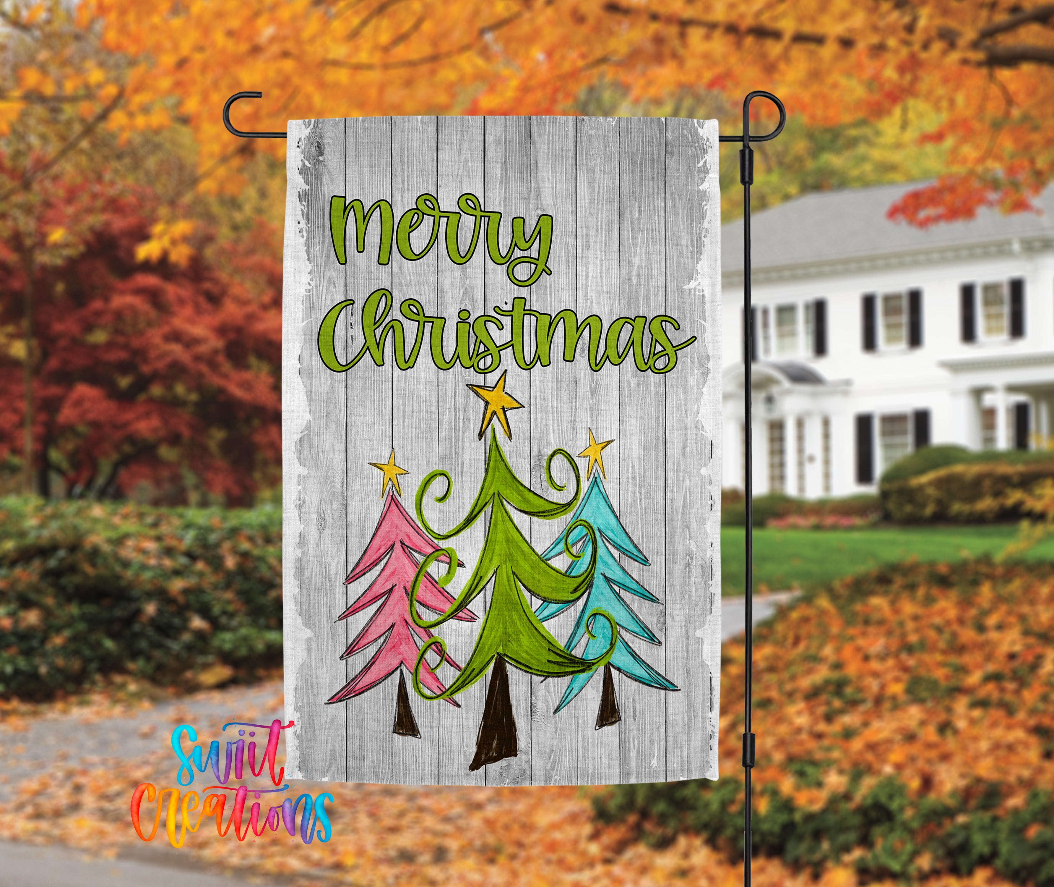 A wooden flag with a festive Christmas design hangs in front of a white house with autumn foliage in the background.