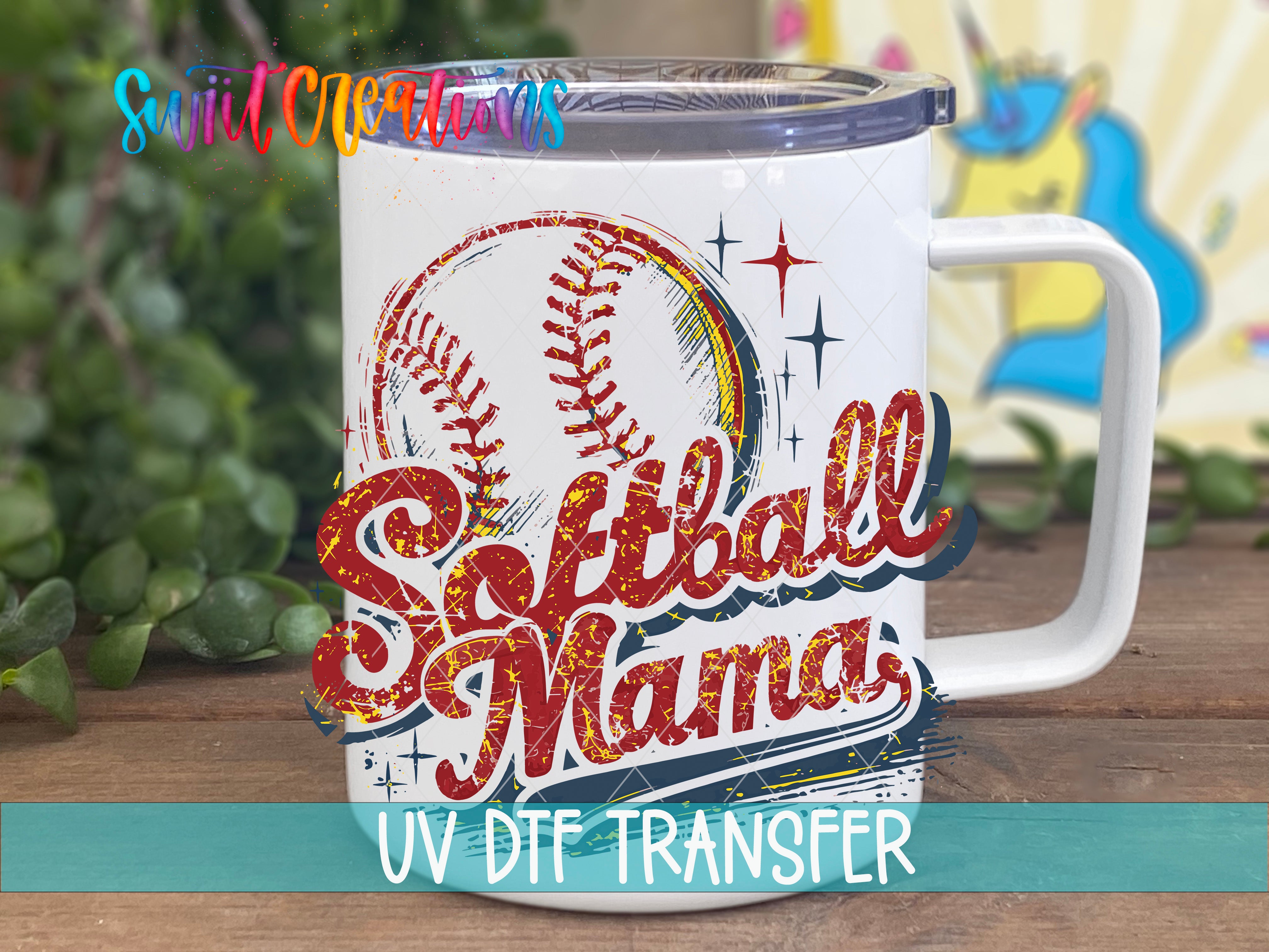 A white mug with a baseball and softball design, along with the text "Softball Mama" printed on it. The mug is placed on a wooden surface, and there is a blurred background that includes some greenery.